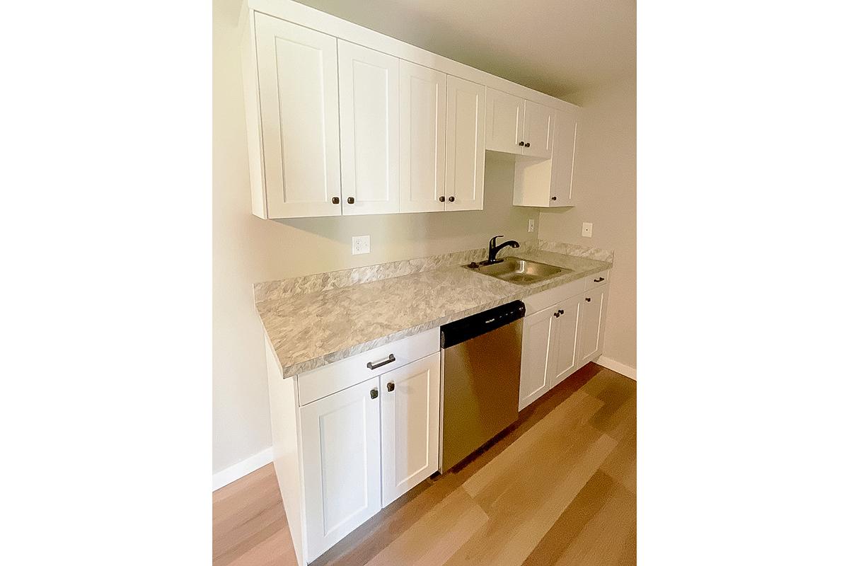 A modern kitchen featuring white cabinetry, a stainless steel dishwasher, a sink with a black faucet, and a light-colored countertop. The kitchen has a minimalist design with a neutral wall color and wooden flooring, providing a clean and spacious appearance.