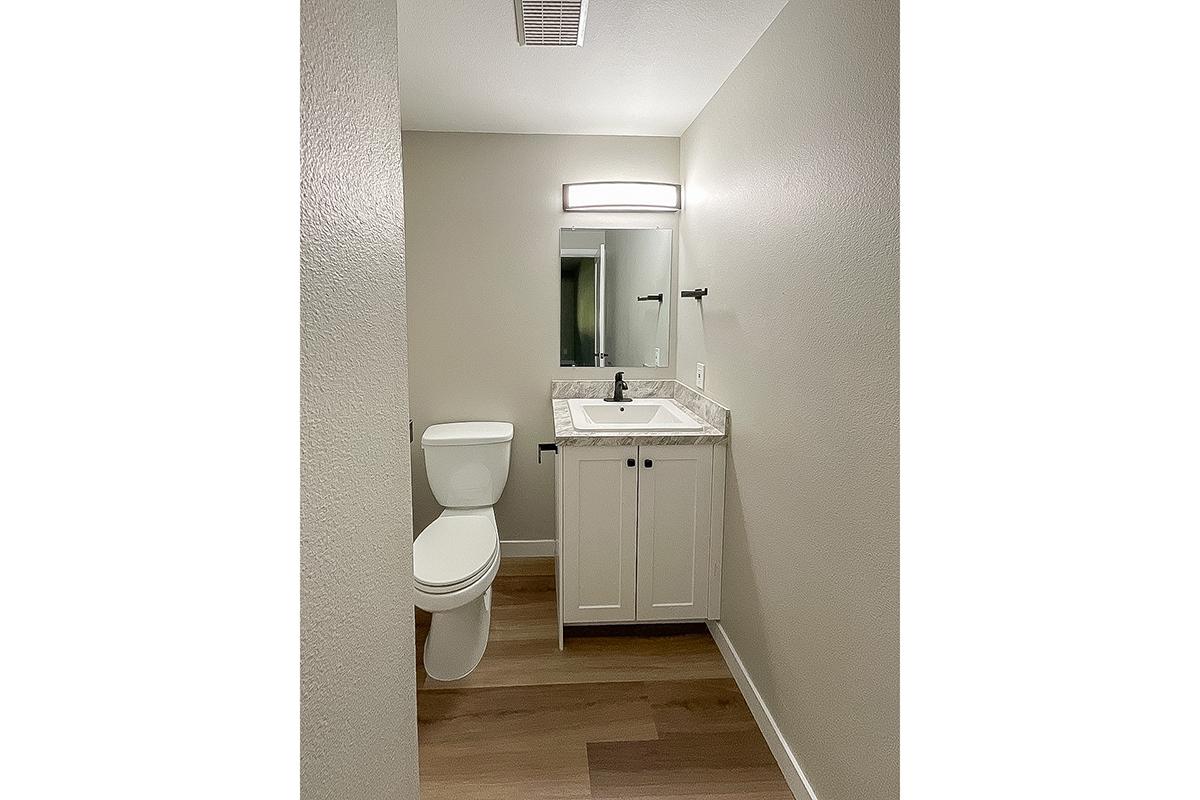 Modern bathroom featuring a white toilet and vanity with a sink. The vanity has a marble countertop and a mirror above it. Soft overhead lighting illuminates the space, and light-colored walls complement the wood flooring. The overall design is clean and minimalist.