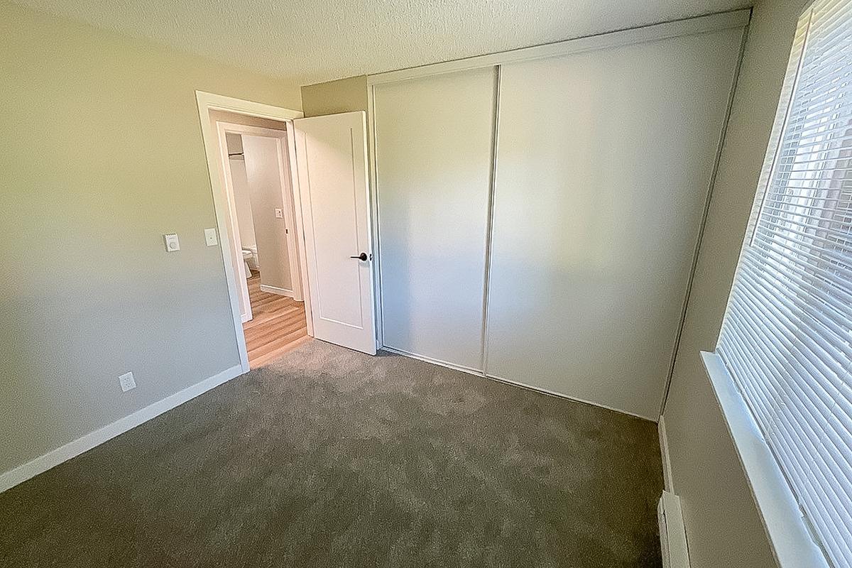 A well-lit interior room with light gray walls and carpet flooring. There is a closed sliding door on the left, leading to a closet, and an open door in the background that appears to lead to a bathroom. The room has a window with blinds allowing natural light to enter.