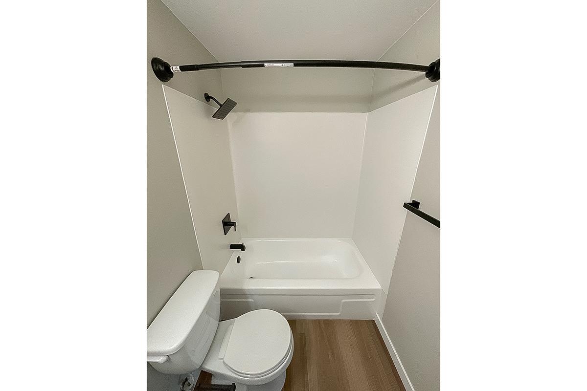 A small bathroom featuring a bathtub with a black shower curtain rod, a wall-mounted shower head, a toilet, and light-colored walls. The floor is made of light wood, and there are minimal fixtures, creating a clean and modern look.