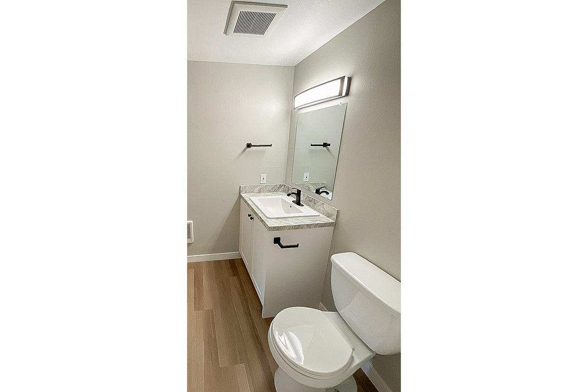 A modern bathroom featuring a white toilet and a sink with a marble countertop. Above the sink is a mirror and a sleek light fixture. The walls are painted in a neutral color, and the floor has a wood-like appearance. There are towel bars mounted on the wall.
