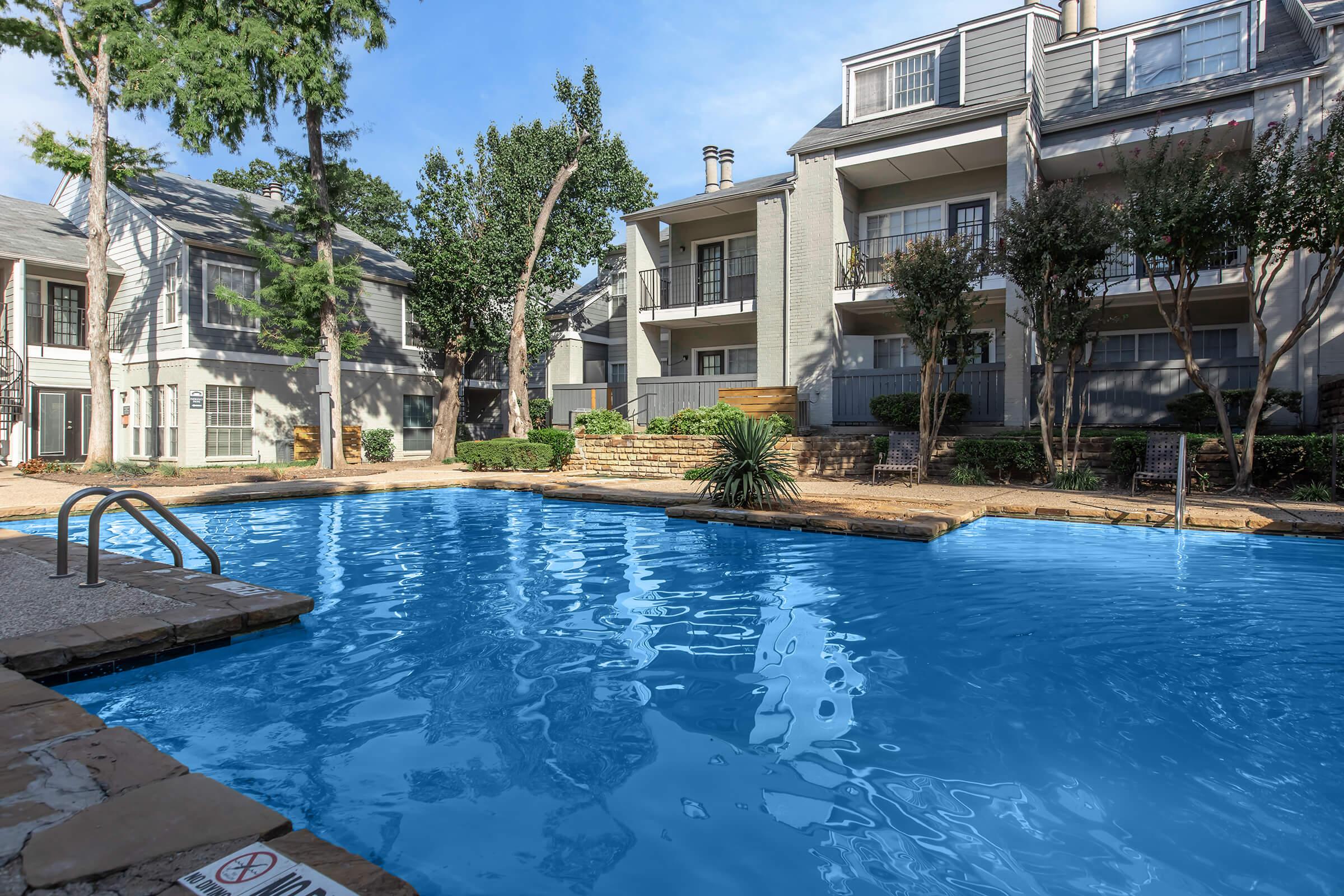 A clear blue swimming pool surrounded by stone and concrete, with lush greenery and trees nearby. Multi-story residential buildings with balconies are visible in the background. Sunlight reflects off the water, creating a warm and inviting atmosphere.