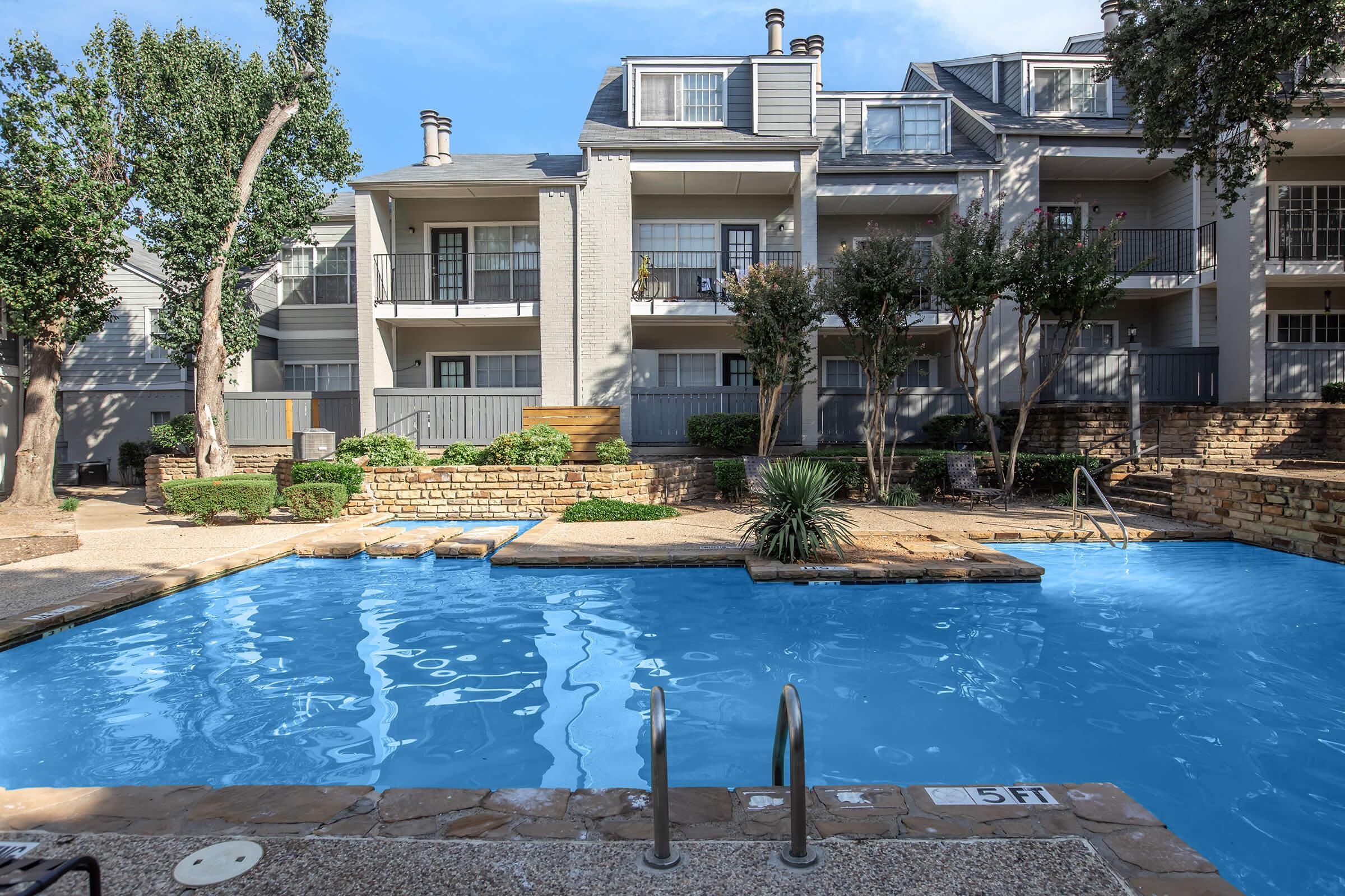 Swimming pool in an apartment complex, featuring clear blue water and surrounding landscaped areas with trees and shrubs. In the background, several floors of the residential building are visible, with balconies and patio spaces. Sunlight illuminates the scene, creating a serene atmosphere.