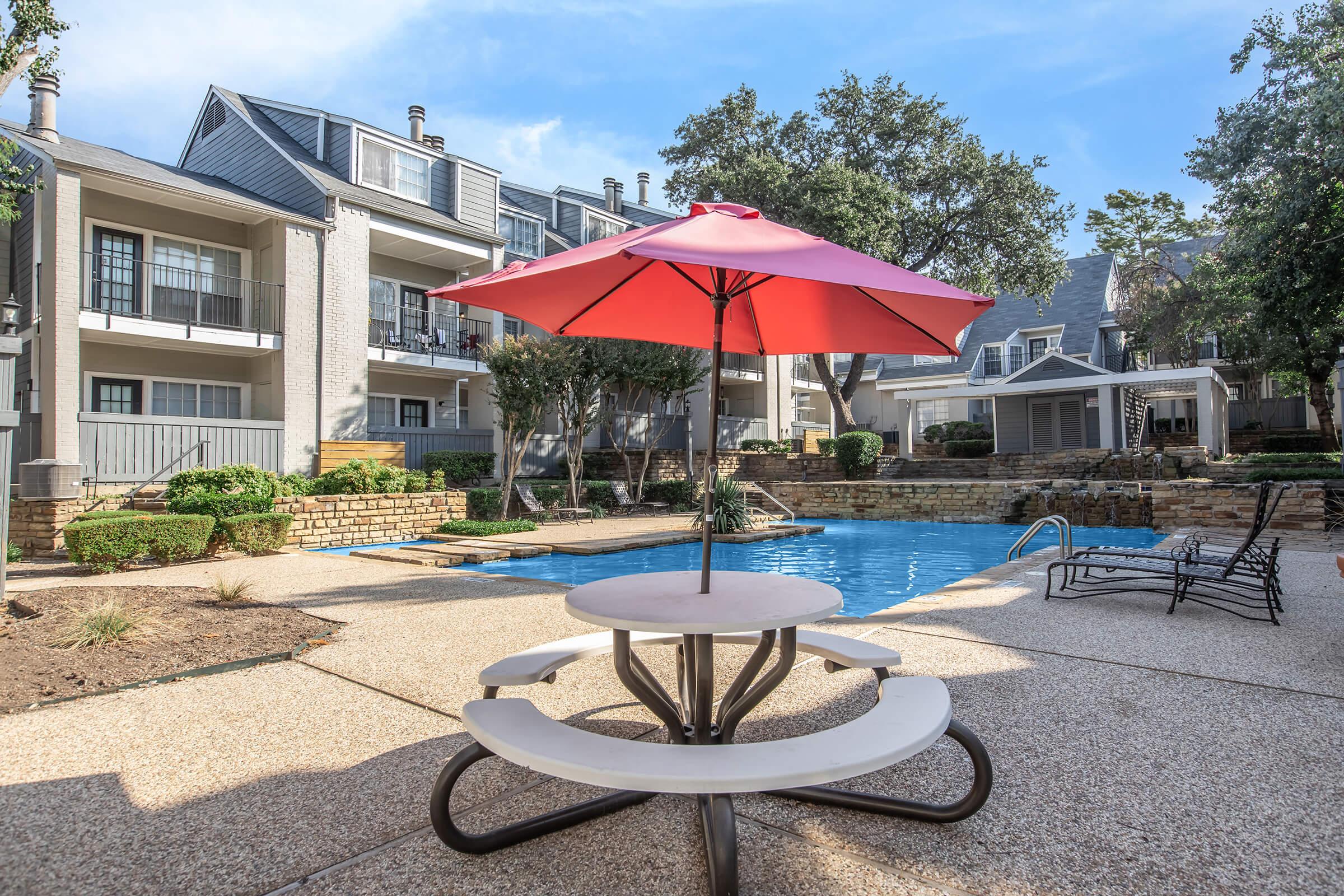A swimming pool area with a circular table and benches under a red umbrella. Surrounding the pool are landscaped gardens and a few lounge chairs. In the background, there are multi-story residential buildings with balconies and trees. The sky is clear and sunny, creating a relaxing atmosphere.