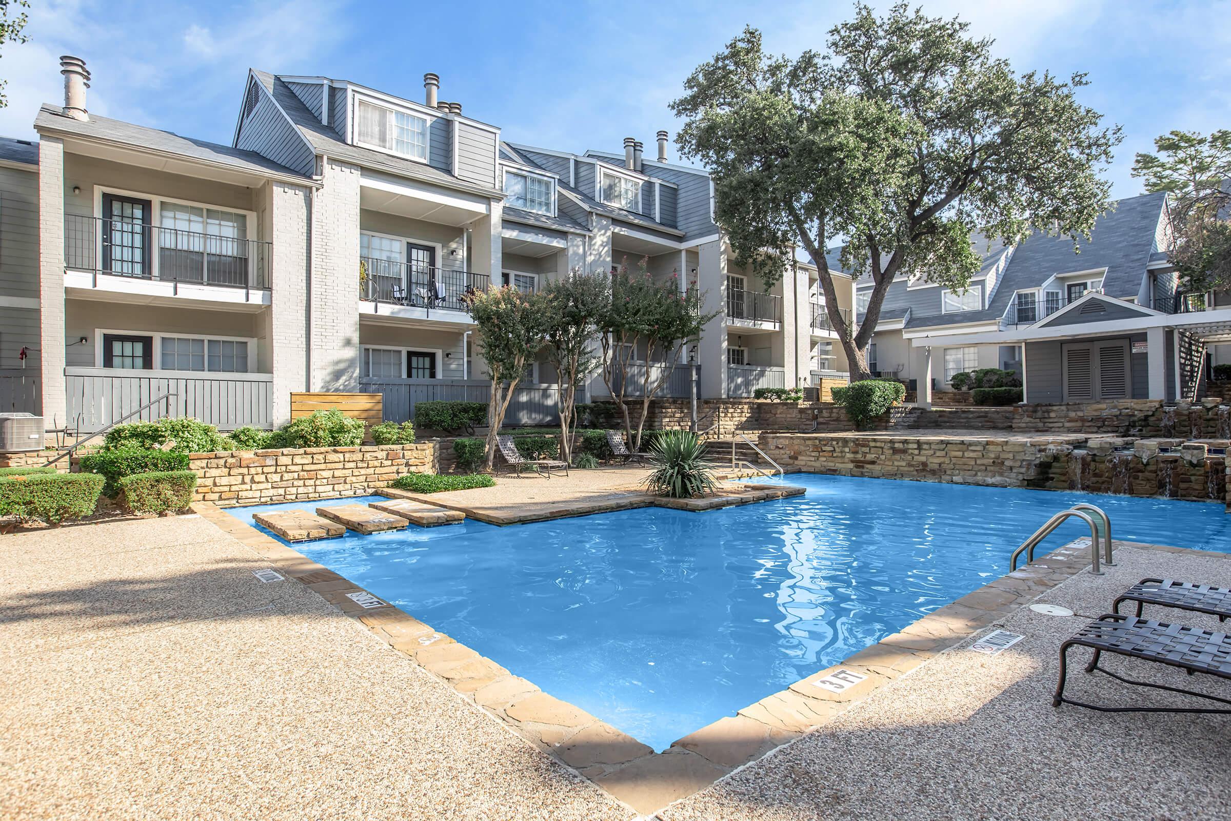 A serene outdoor swimming pool surrounded by landscaped greenery and residential buildings. The pool features a stone border, lounge chairs nearby, and is set against a clear blue sky, creating a relaxing atmosphere.