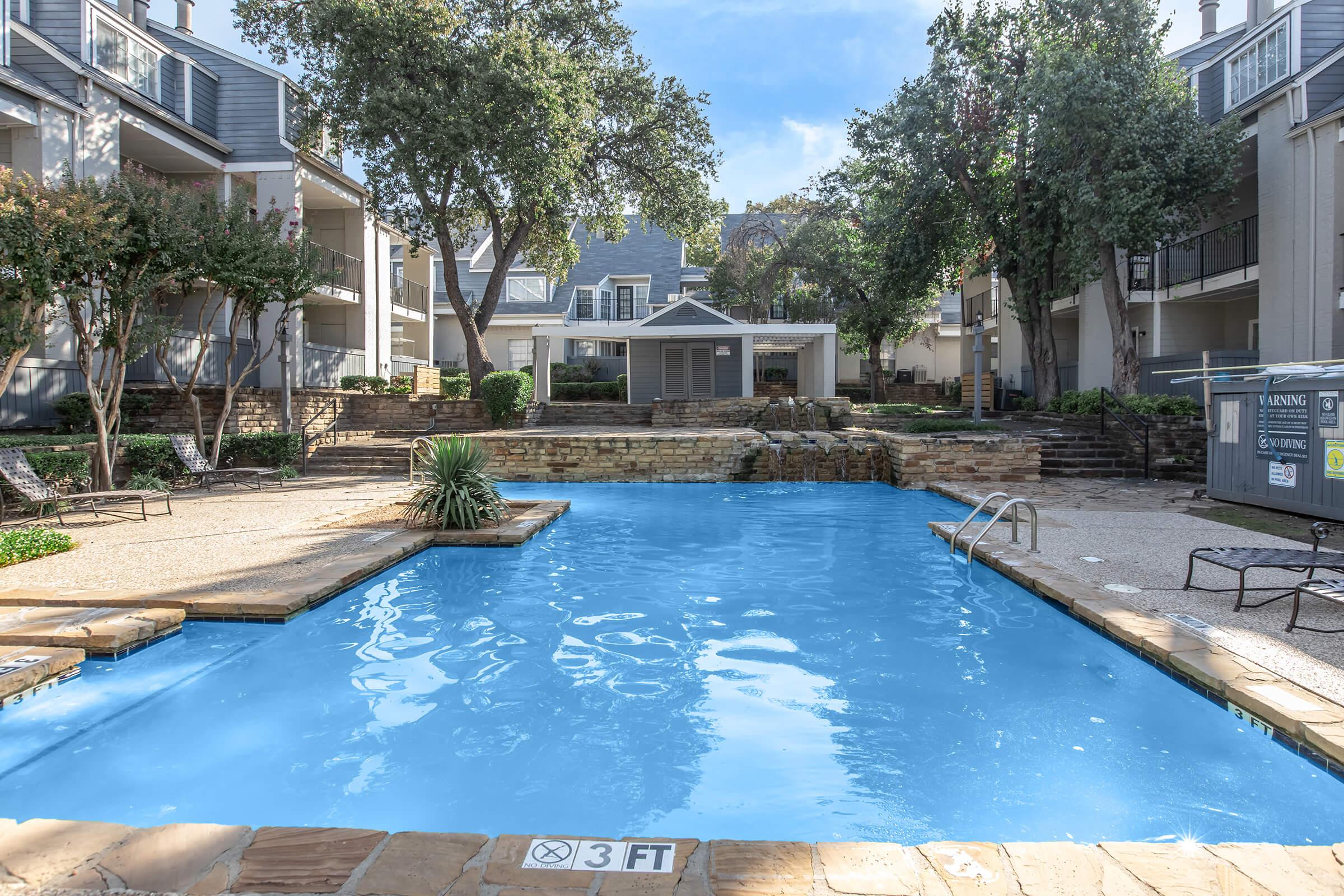 A bright outdoor swimming pool surrounded by well-maintained landscaping in an apartment complex. The pool features steps leading in, lounge chairs nearby, and a shallow area marked with a depth sign. Trees and buildings frame the scene under a clear blue sky.