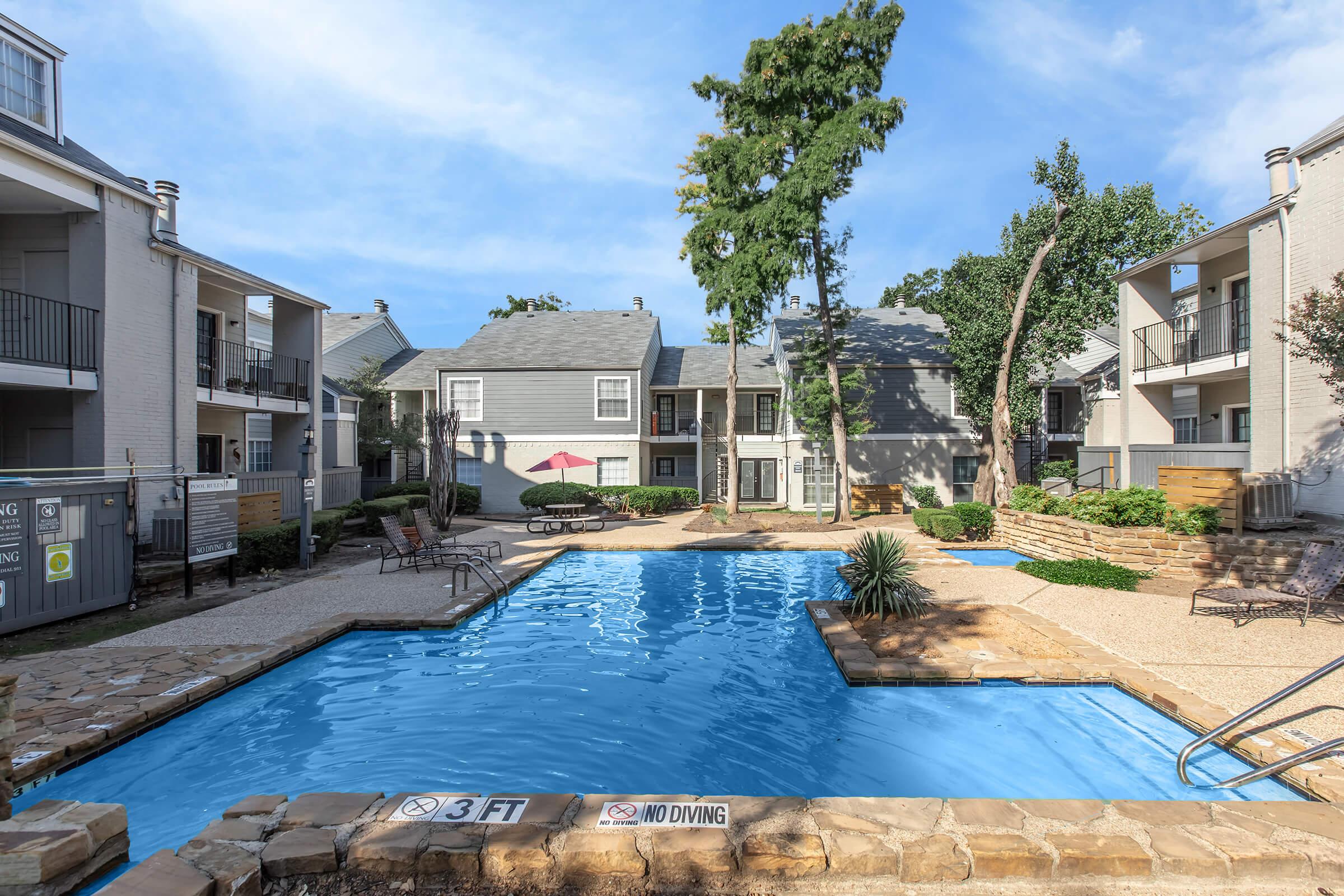A clear blue swimming pool surrounded by landscaped areas and apartment buildings. The pool has a shallow end marked with a "3 FT" sign, and there is a "NO DIVING" warning. Lounge chairs are positioned nearby, and trees are dotted around the area under a bright blue sky.