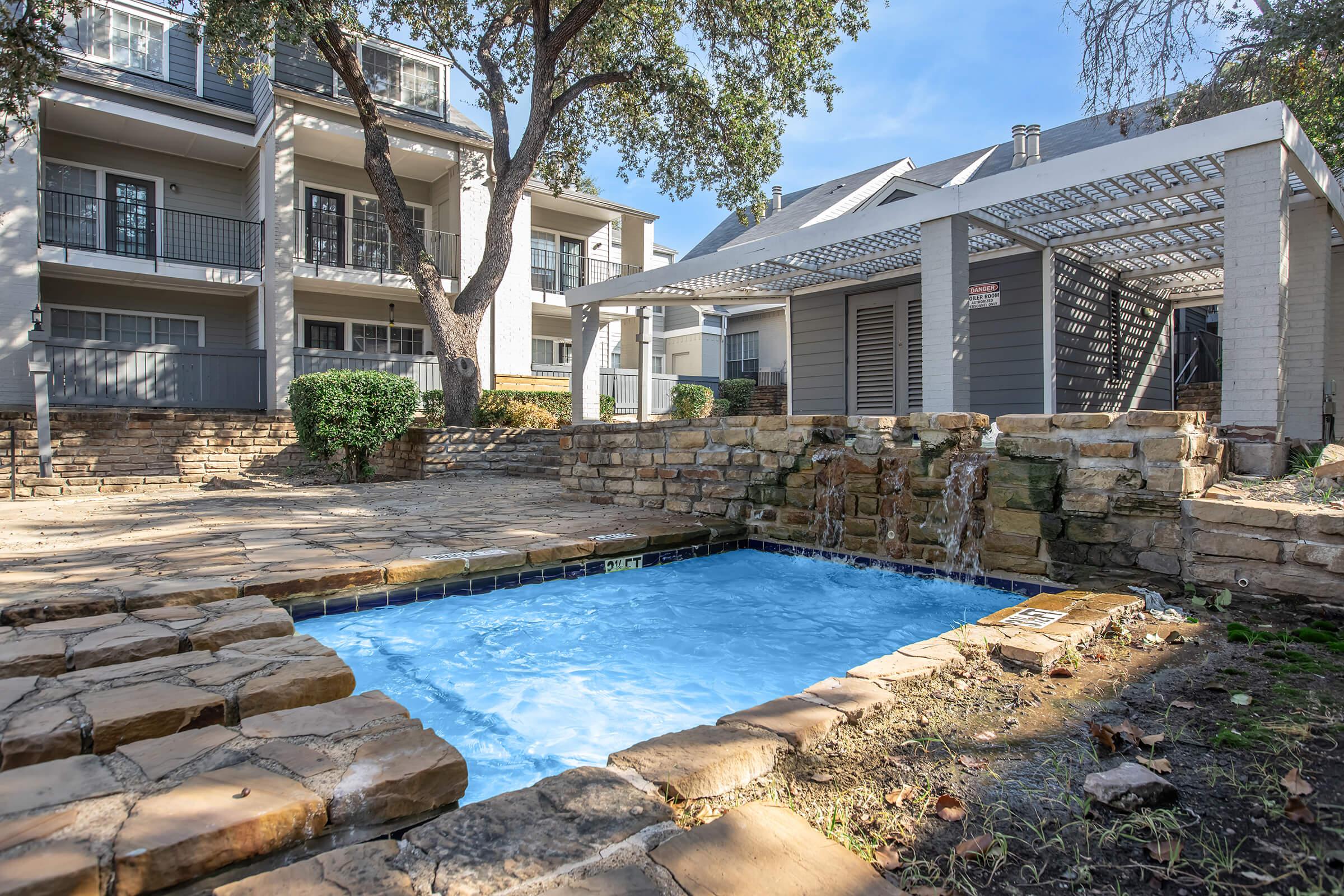 A serene outdoor area featuring a small swimming pool with clear blue water, surrounded by natural stone landscaping. In the background, there are multi-story apartment buildings and a covered patio with a trellis. Lush green trees provide shade, creating a relaxing atmosphere.