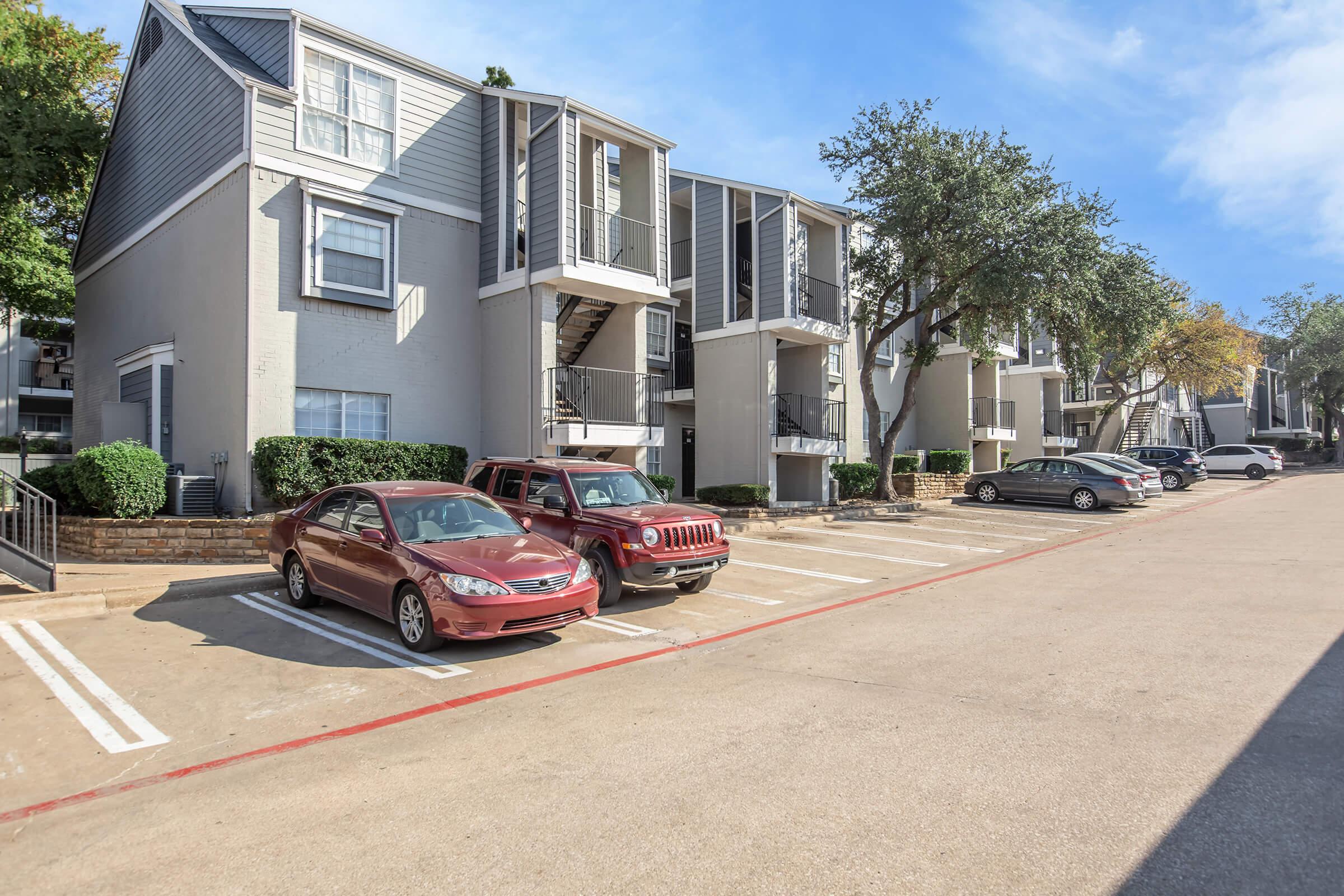 Exterior view of an apartment complex featuring two buildings with multiple balconies. The parking lot has several cars, including a red sedan and a red SUV. Trees and landscaping surround the area, providing a pleasant environment. Clear blue sky overhead.