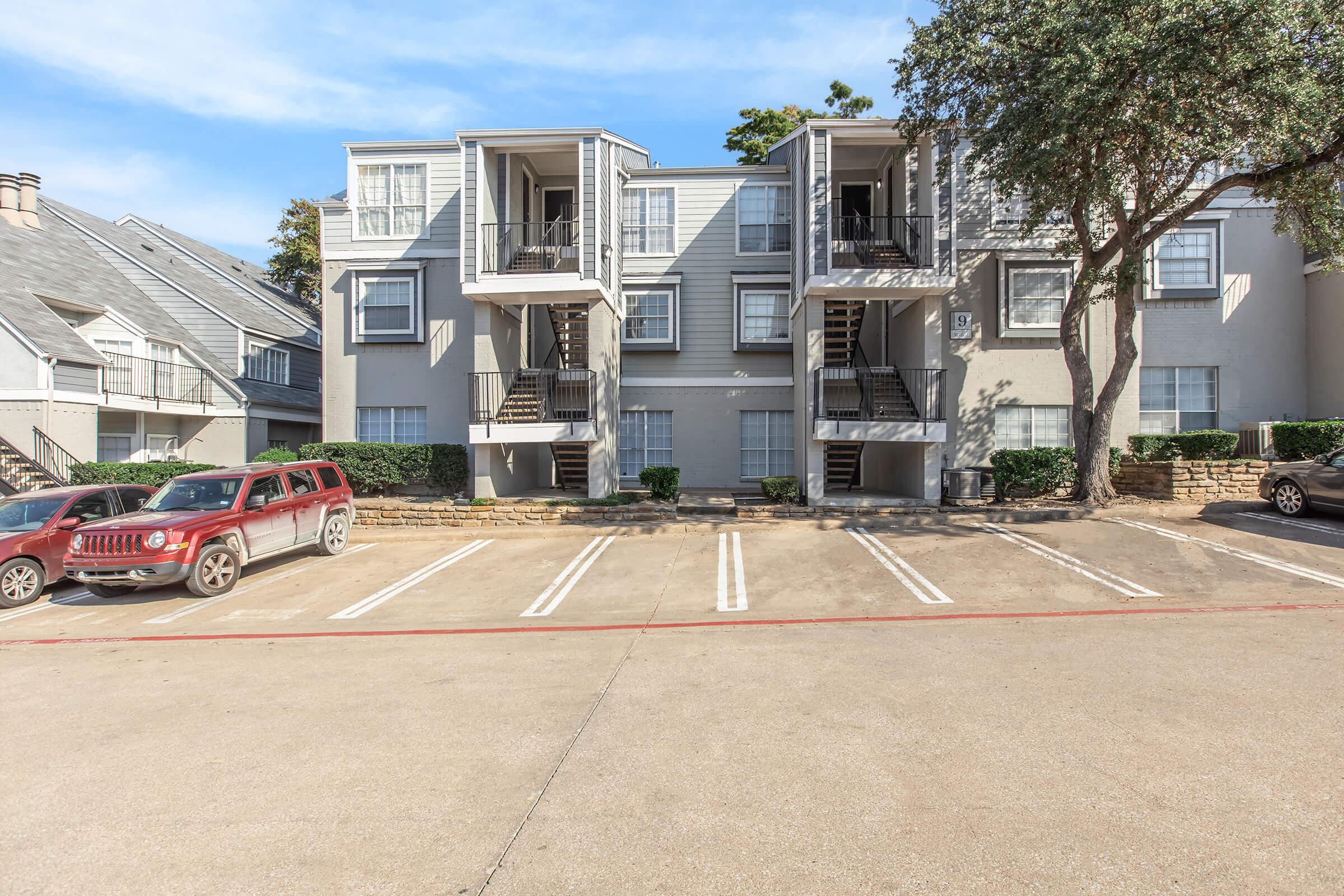 A multi-unit residential building with multiple balconies, surrounded by greenery. In the foreground, a parking lot with several parked cars, including a red SUV. The scene is set under a clear blue sky, highlighting the architectural details of the building and the arranged parking spaces.