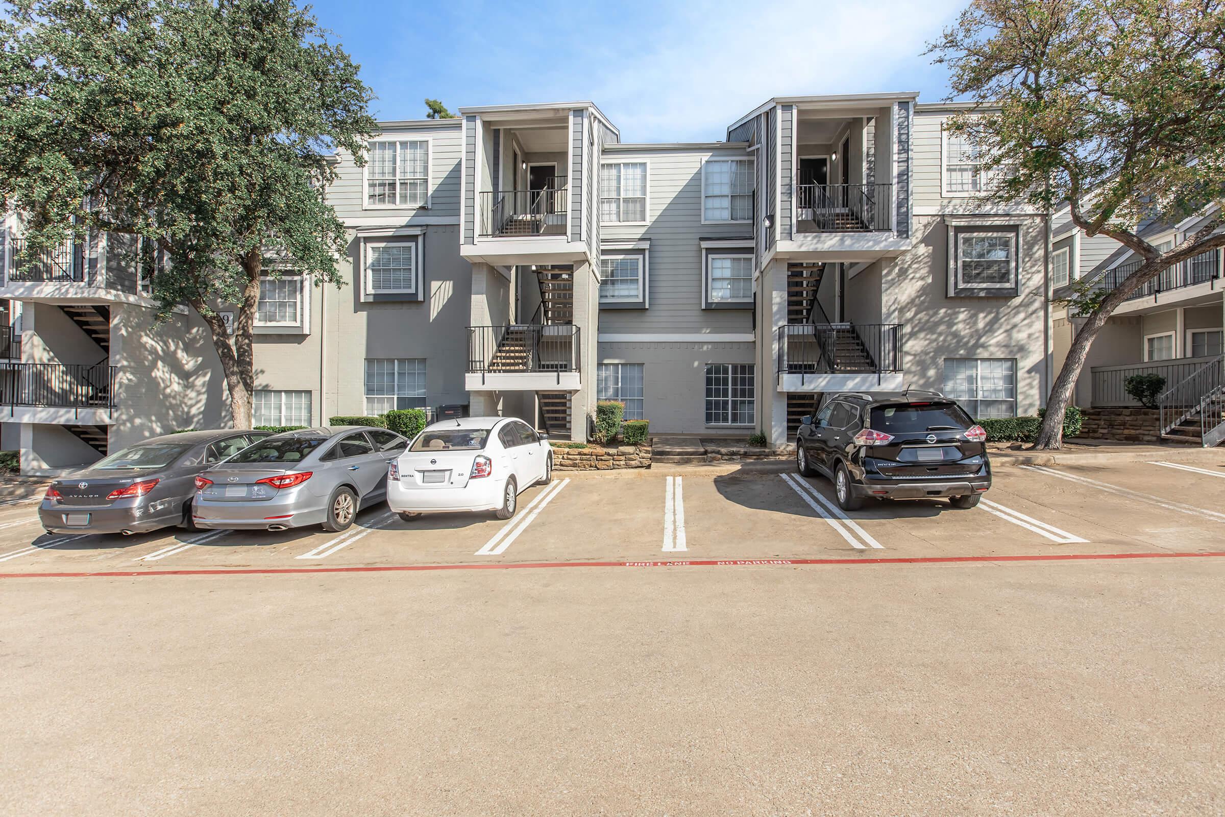 A multi-unit apartment complex featuring two-story buildings with staircase access. The building has a light gray exterior and large windows. In the foreground, several cars are parked in designated spaces, with a tree providing shade to one side. The sky is clear, indicating a sunny day.