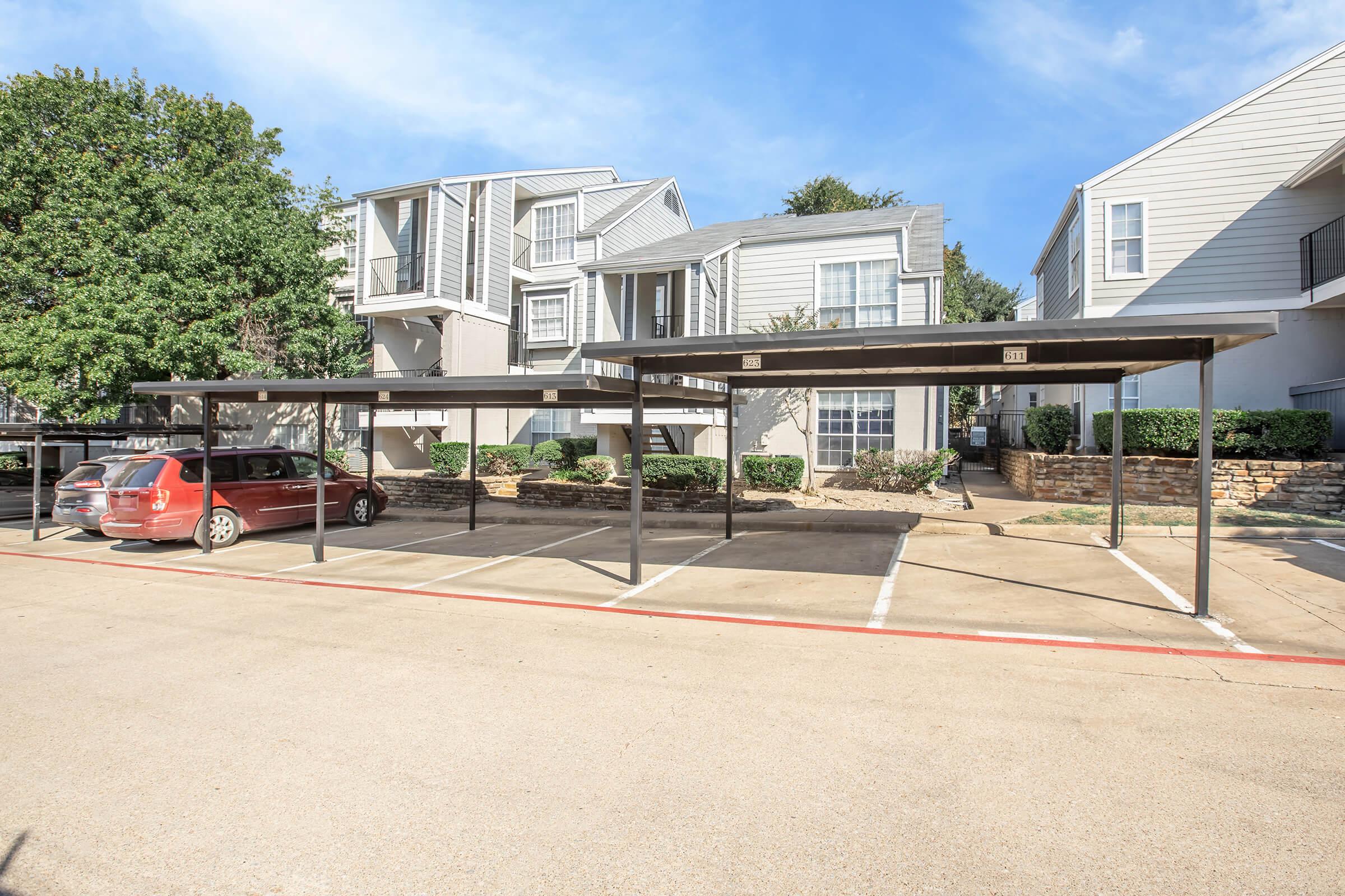A parking lot with a few covered parking spaces in front of two apartment buildings. There is one red car parked in a space, and the area is surrounded by greenery and landscaping. The sky is clear and blue, indicating a sunny day.