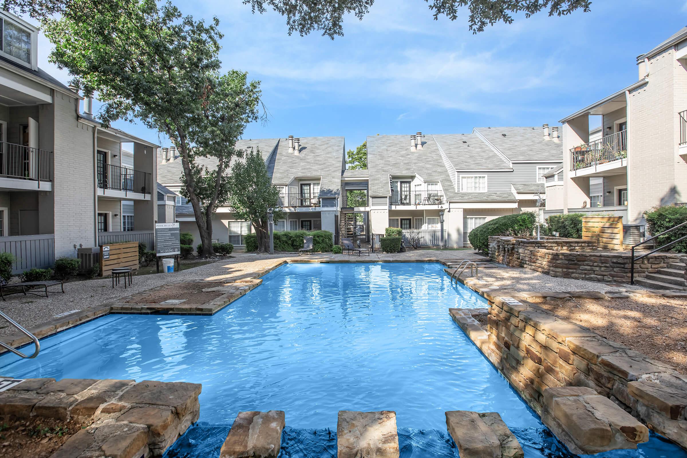 A tranquil apartment complex featuring a swimming pool surrounded by landscaped greenery. The pool is clear and inviting, with stone pathways leading into it. Nearby are residential buildings with balconies, set against a clear blue sky. The atmosphere is peaceful, ideal for relaxation.