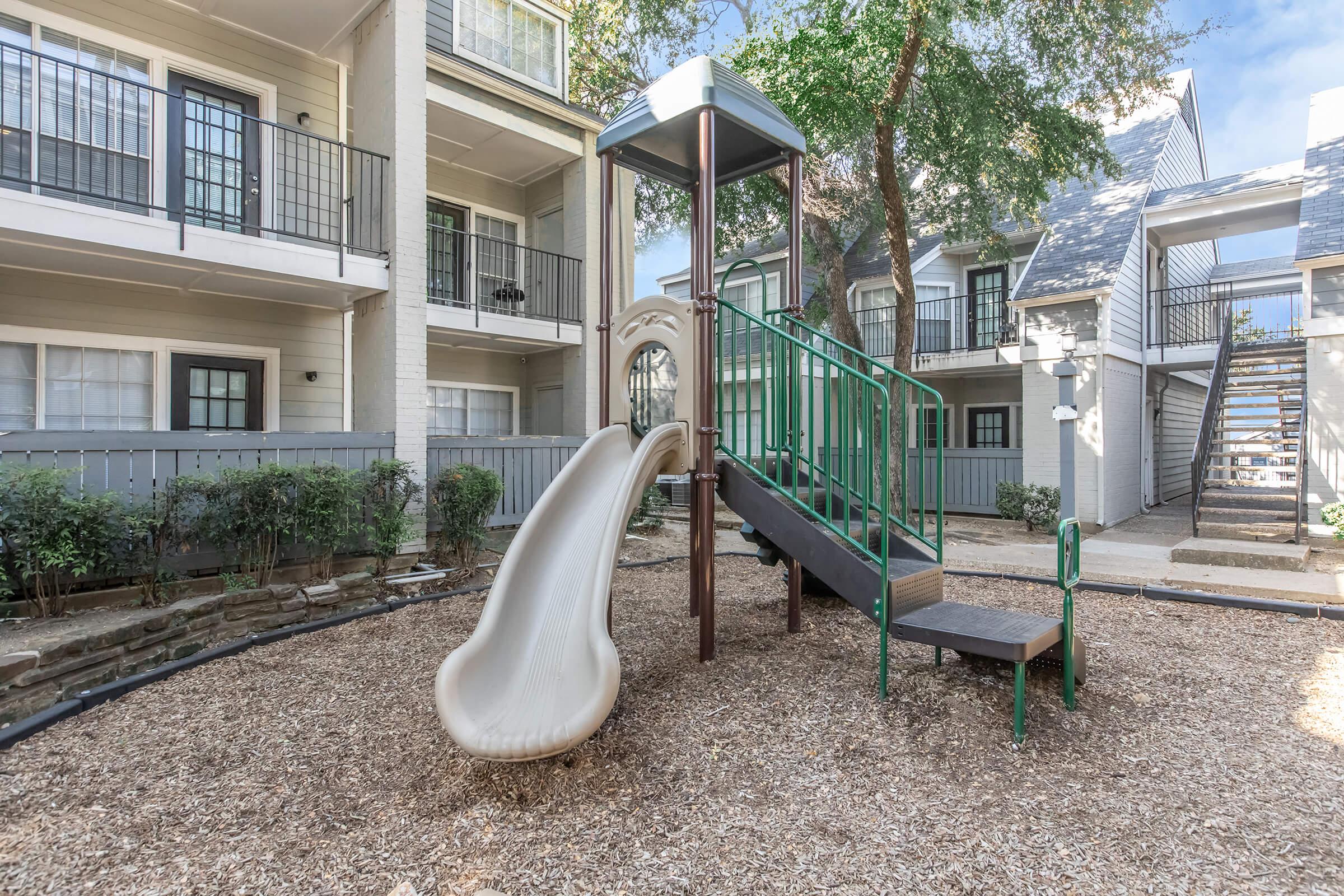 A small playground featuring a slide, climbing structure, and a bench, surrounded by an area of wood chips. The playground is situated in a residential complex, with two-story buildings in the background and greenery along the edges. Sunny blue sky above.
