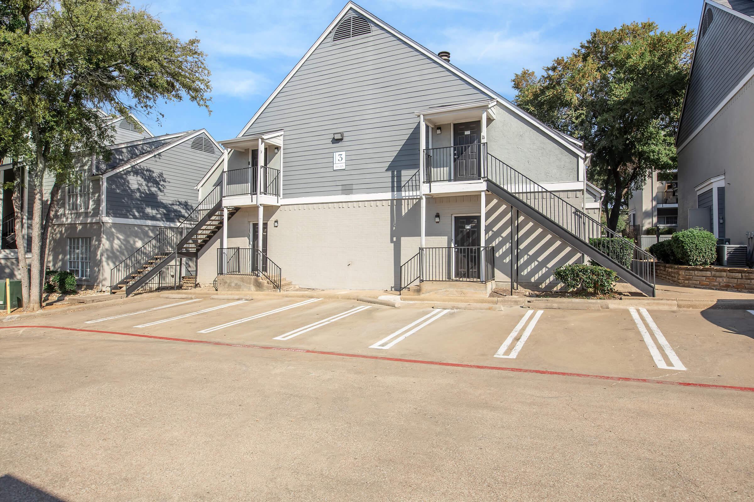 Two-story residential building with gray siding, featuring a stairway entrance. Surrounding area includes a parking lot with marked spaces and landscaping with trees and bushes. Clear blue sky and well-maintained exterior.