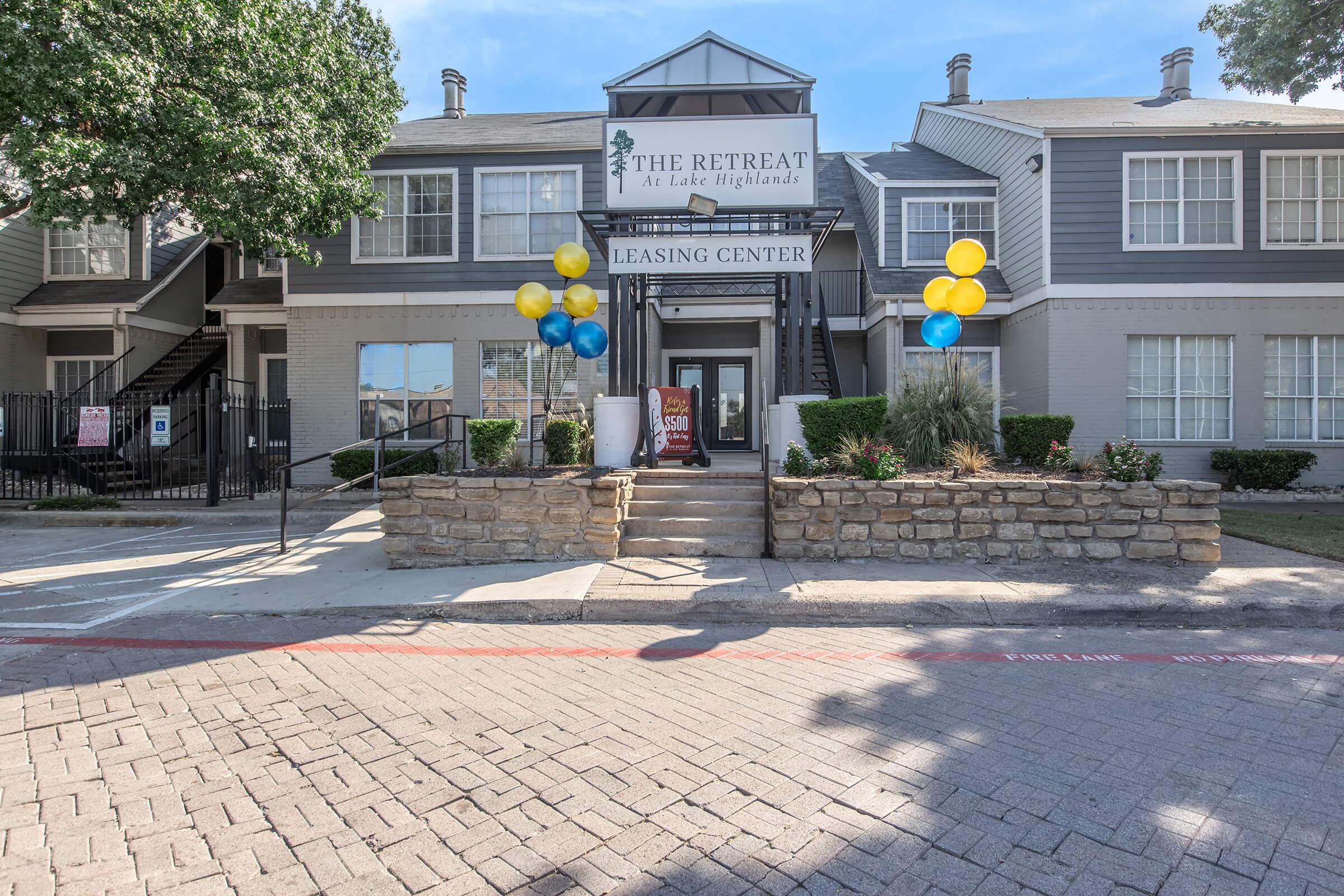 Leasing center entrance of "The Retreat at Lake Highlands," featuring a stone pathway, decorative balloons in blue and yellow, and a landscaped area with shrubs. The building has large windows and a covered porch, creating an inviting atmosphere.