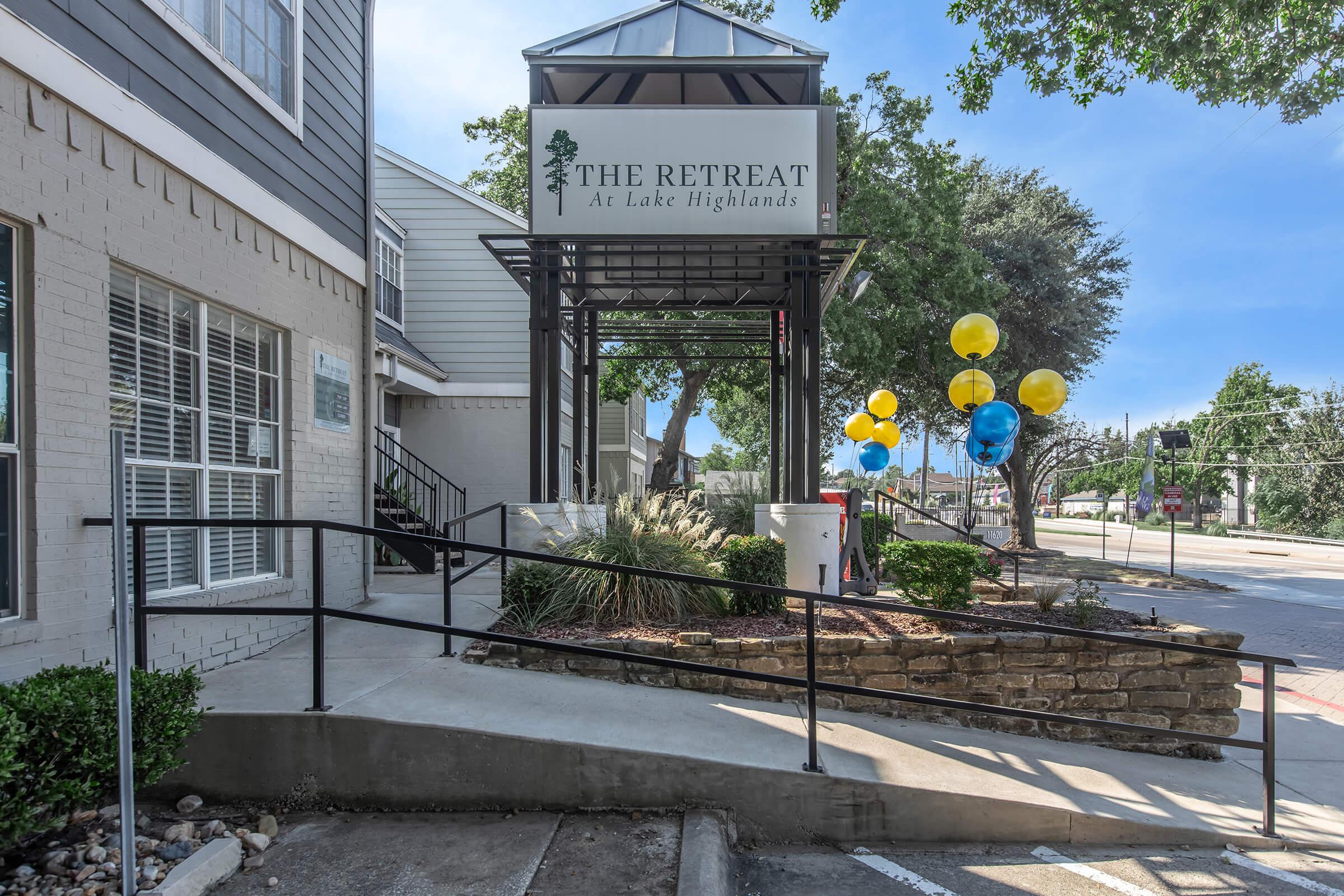 A sign for "The Retreat at Lake Highlands" displayed on a modern building, surrounded by greenery. A ramp leads to the entrance, and colorful balloons in yellow and blue are positioned nearby, adding a festive touch to the scene. The sky is clear and bright, contributing to a welcoming atmosphere.