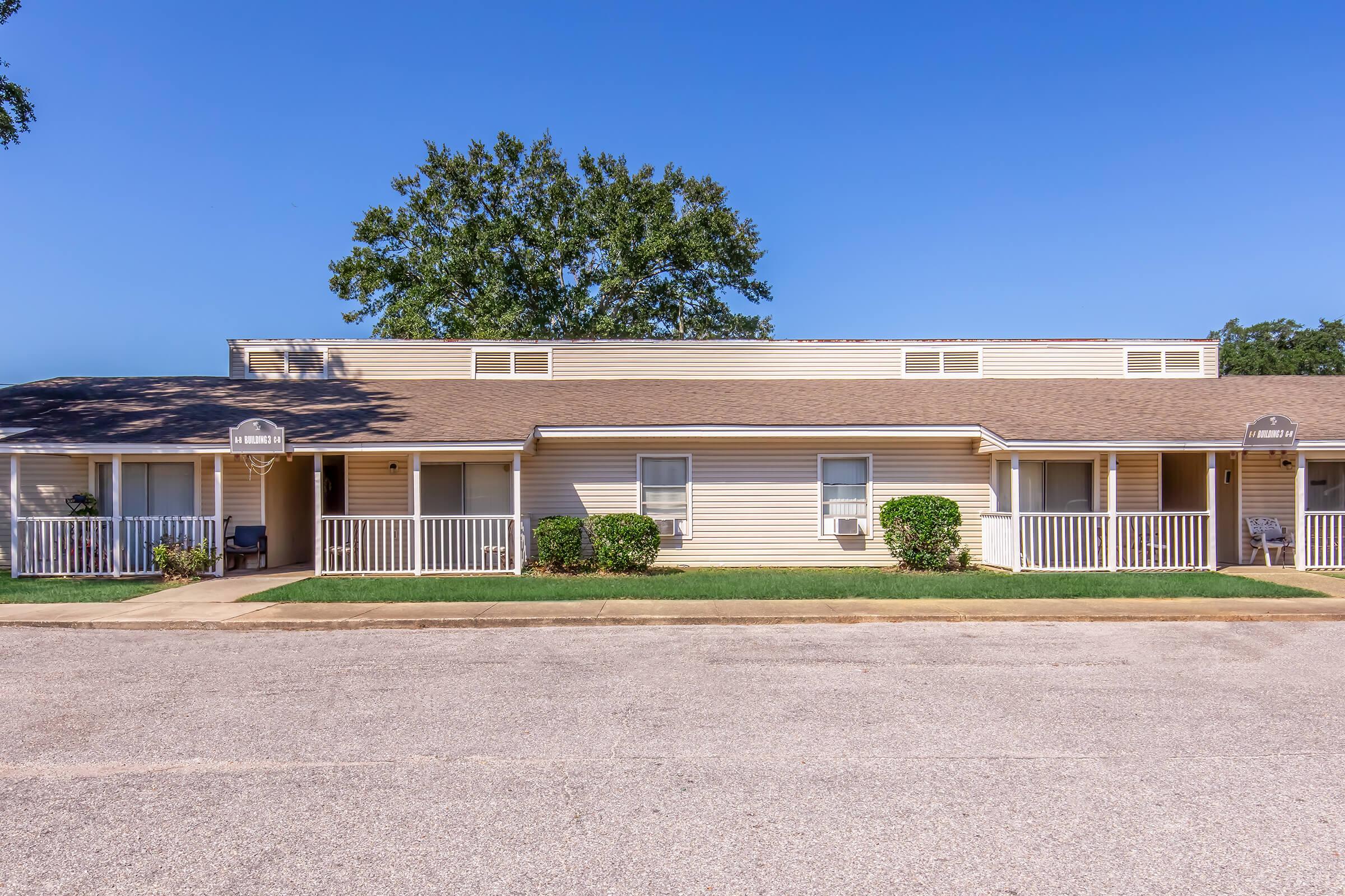 A row of yellowish apartment buildings with white porches, situated on a quiet street. Green grass and small bushes line the path in front. The sky is clear and blue, contributing to a pleasant, sunny atmosphere.