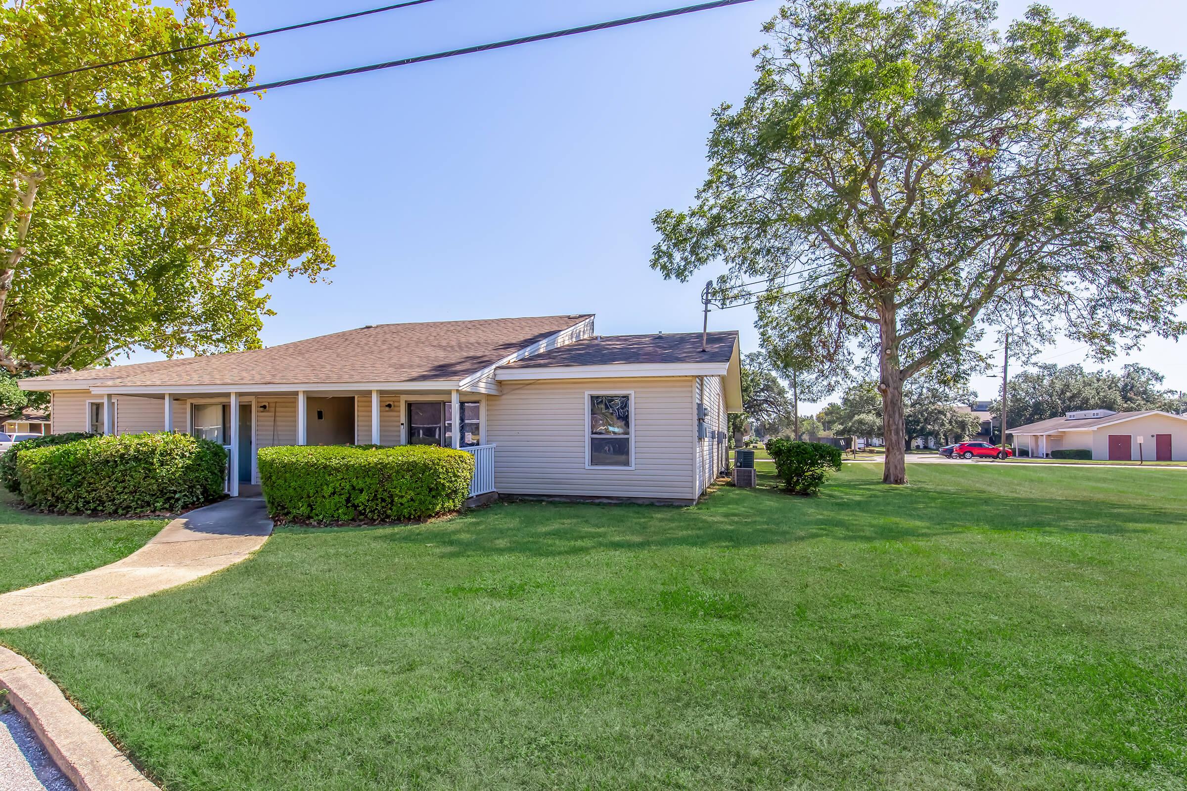 A well-maintained single-story building featuring a landscaped front yard with green grass and shrubs. A concrete walkway leads to the entrance. In the background, a large tree provides shade, and additional buildings are visible in the distance under a clear blue sky.