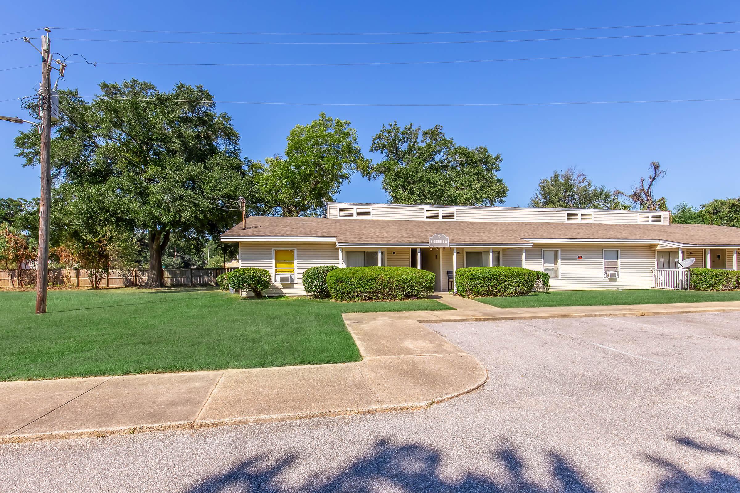 A single-story apartment building with yellow window shutters surrounded by well-maintained green grass and shrubs. Clear blue sky overhead, with a nearby power pole and trees in the background. The parking area in front is paved and smoothly curves towards the building entrance.