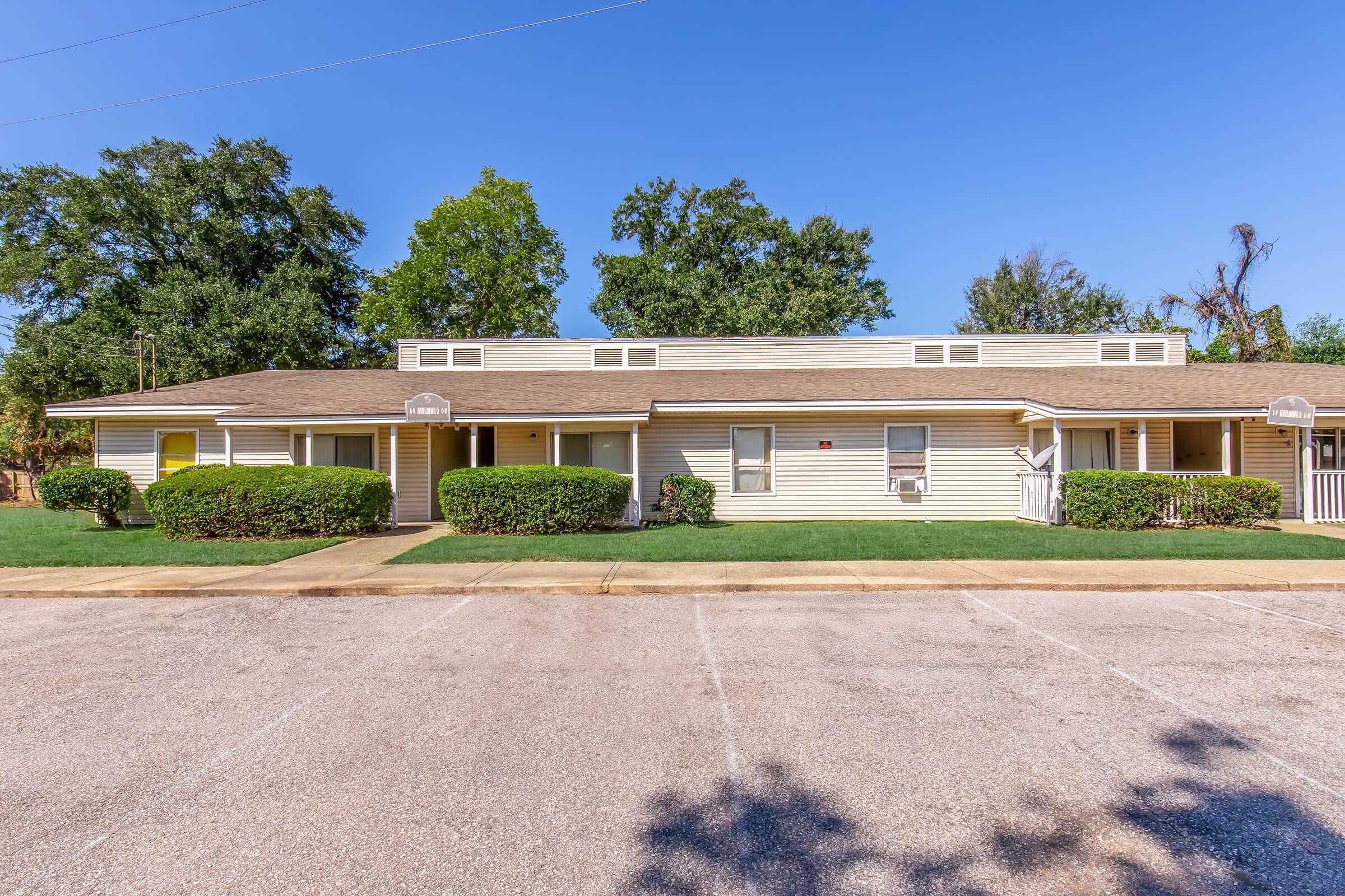 A one-story residential building with multiple units, featuring a light-colored exterior. The structure has a flat roof, several windows, and is surrounded by neatly trimmed bushes and grass. A paved parking area is visible in the foreground, set against a clear blue sky and green trees in the background.