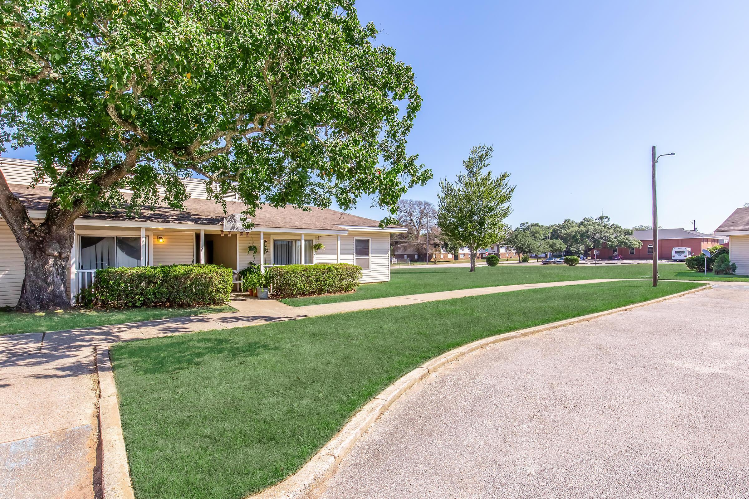 A well-maintained residential area featuring a grassy lawn, a tree, and a sidewalk. The building has a tan exterior with white trim and a covered porch. Clear blue sky overhead enhances the bright, inviting atmosphere.