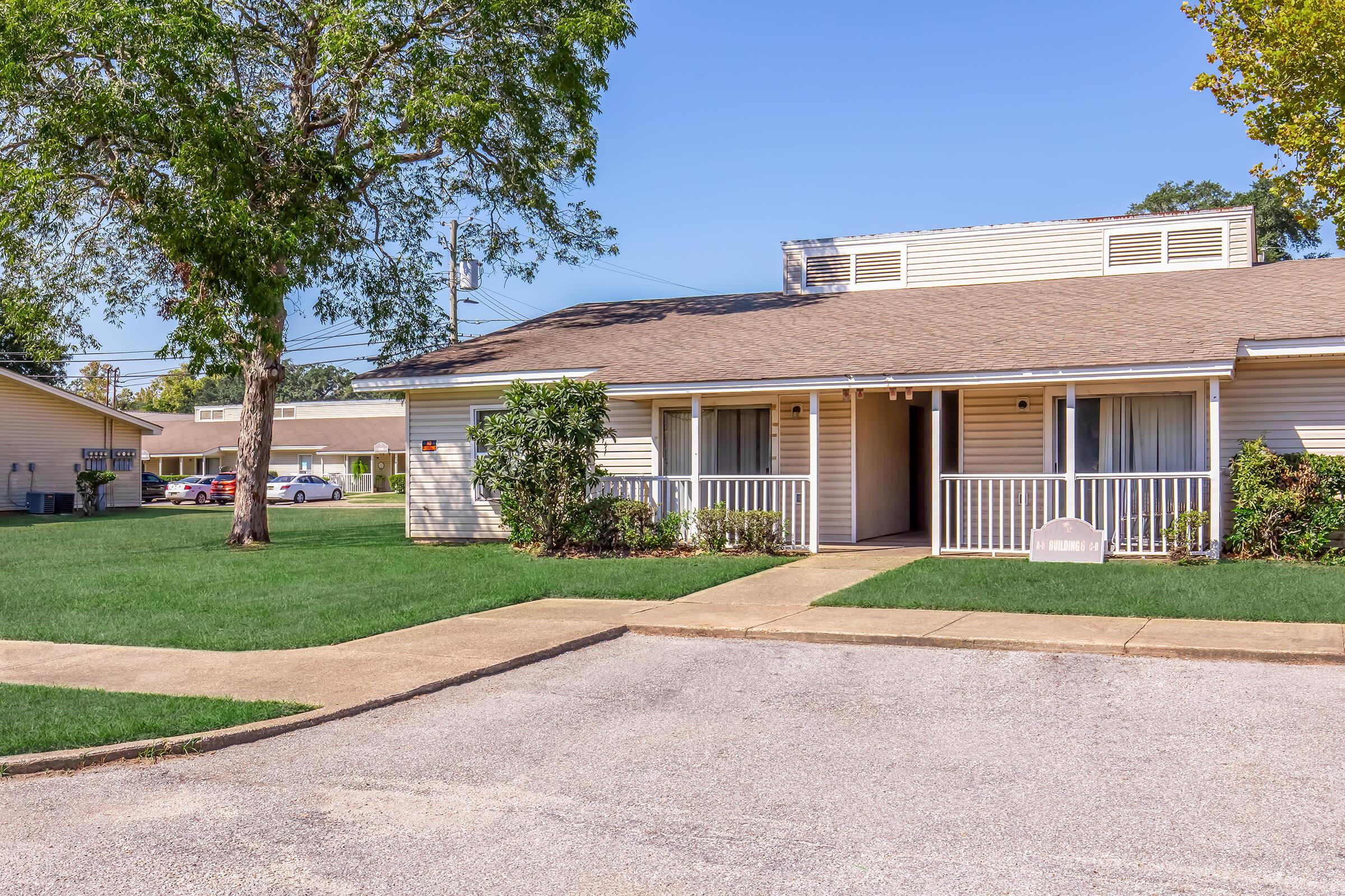 A single-story residential building with a light-colored exterior, featuring a front porch and landscaped greenery. The pathway leads to the entrance, with a well-maintained lawn and nearby parking areas visible in the background under a clear blue sky.