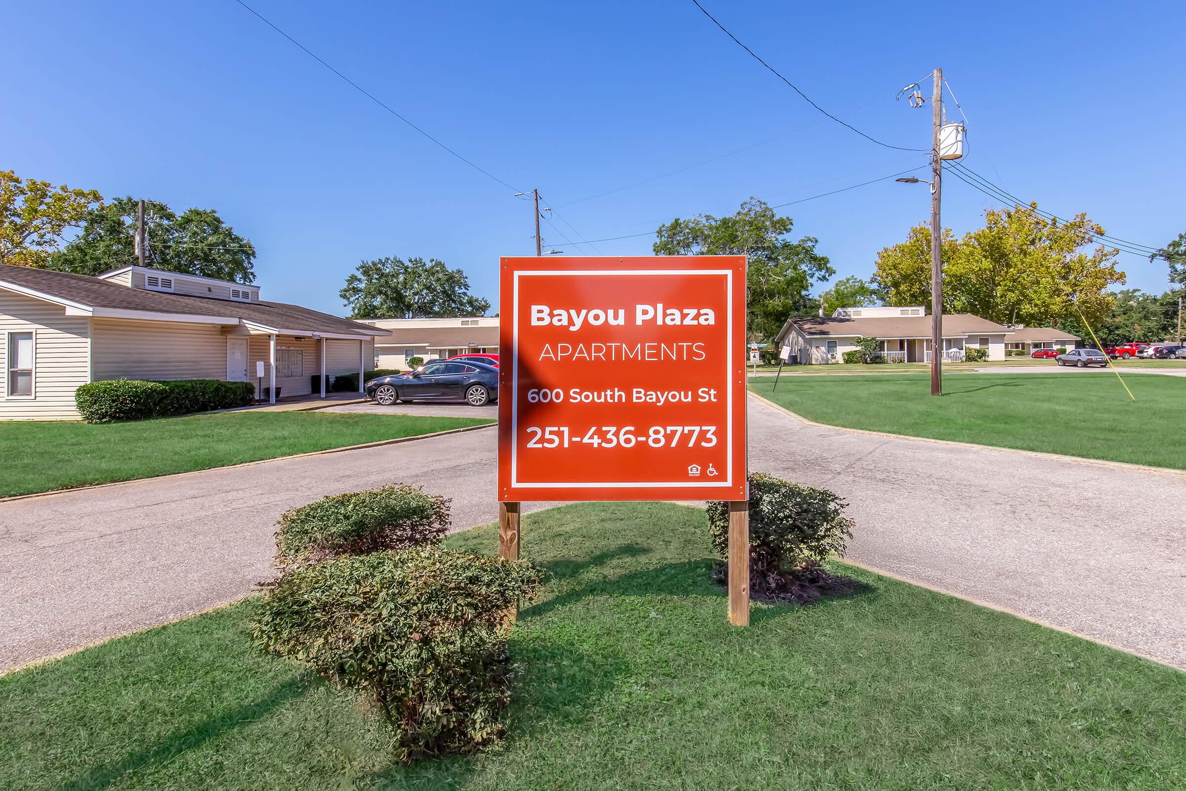 Sign for Bayou Plaza Apartments, located at 600 South Bayou St. The sign displays the contact number 251-436-8773. The setting includes grassy areas and nearby buildings, with a clear blue sky above.