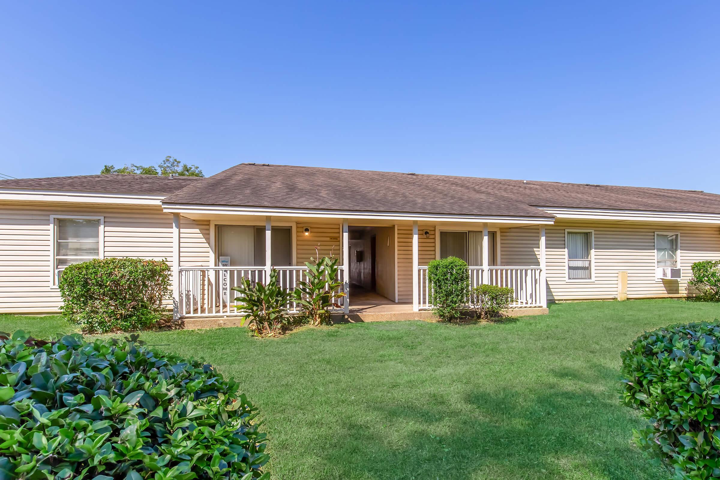 A single-story residential building with a brown roof and light yellow siding. The front features a small porch with white railings, surrounded by well-maintained greenery and lawns under a clear blue sky.