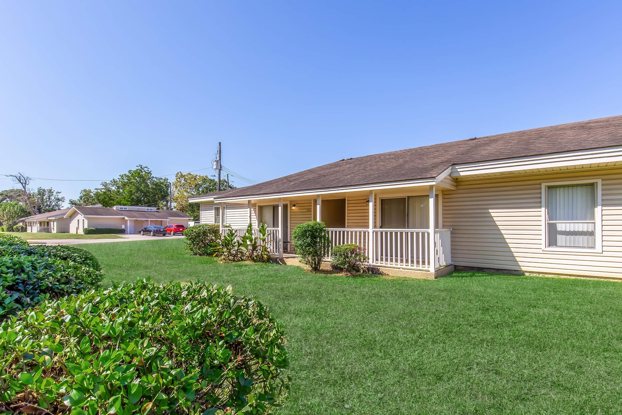 A well-maintained single-story building with a porch, surrounded by neatly trimmed grass and a hedge. In the background, there are additional buildings and trees, indicating a residential area under a clear blue sky.