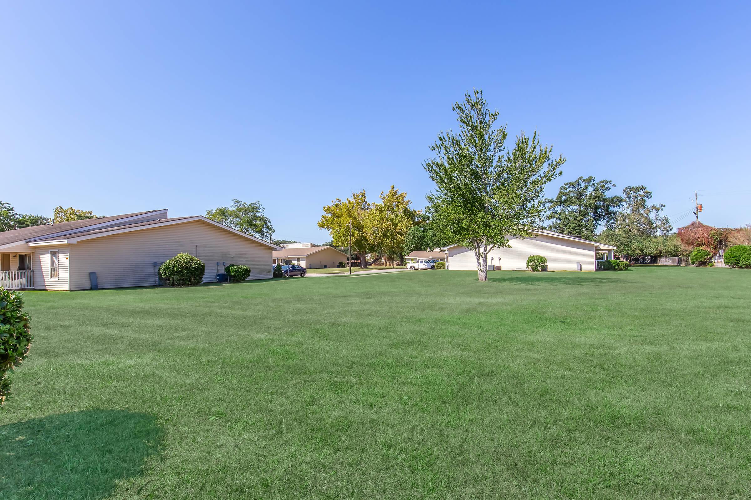 A sunny outdoor scene featuring a green lawn with a few trees. In the background, there are several residential buildings with light-colored exteriors. The sky is clear blue, creating a bright and inviting atmosphere.