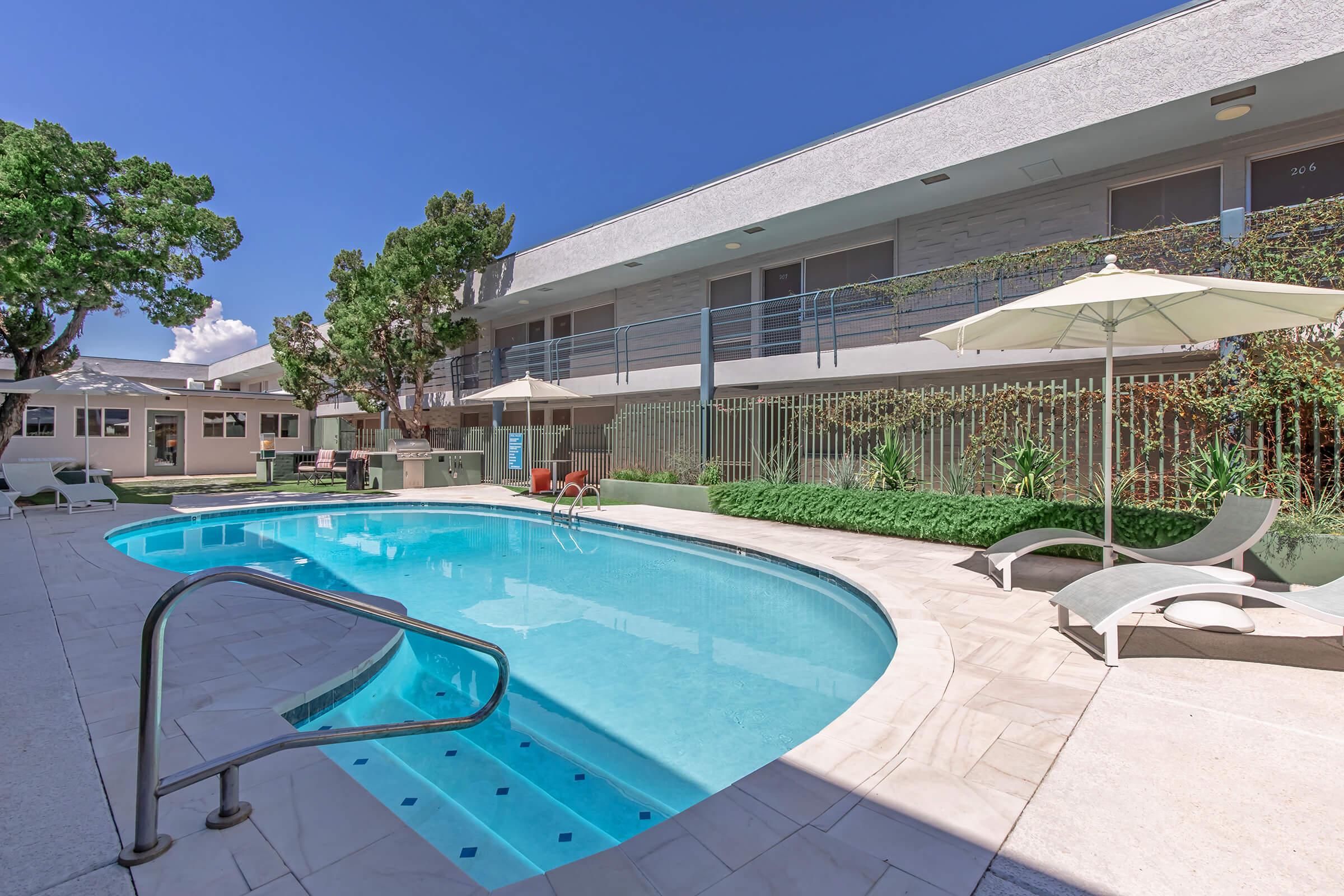 A tranquil outdoor pool surrounded by lounge chairs and umbrellas, set against a modern building with greenery. The clear blue sky enhances the serene atmosphere, making it an inviting space for relaxation and leisure.