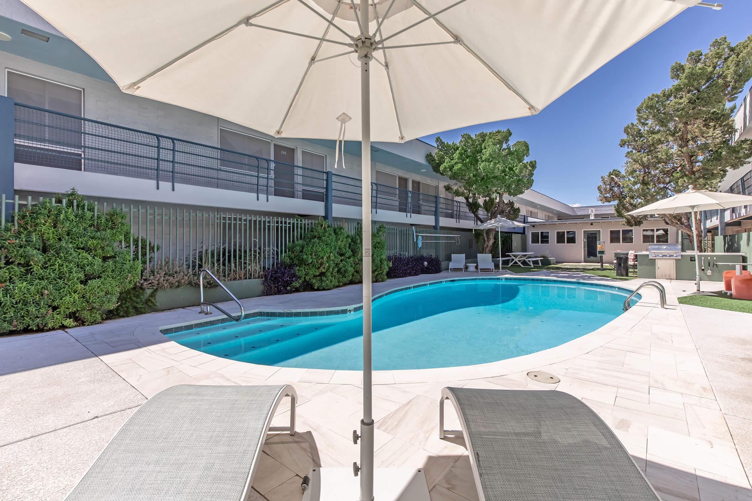 A sunny outdoor pool area with two lounge chairs and a large umbrella in the foreground. The pool is surrounded by greenery and modern buildings. Sunlight casts shadows, creating a relaxing atmosphere perfect for leisure and enjoyment.
