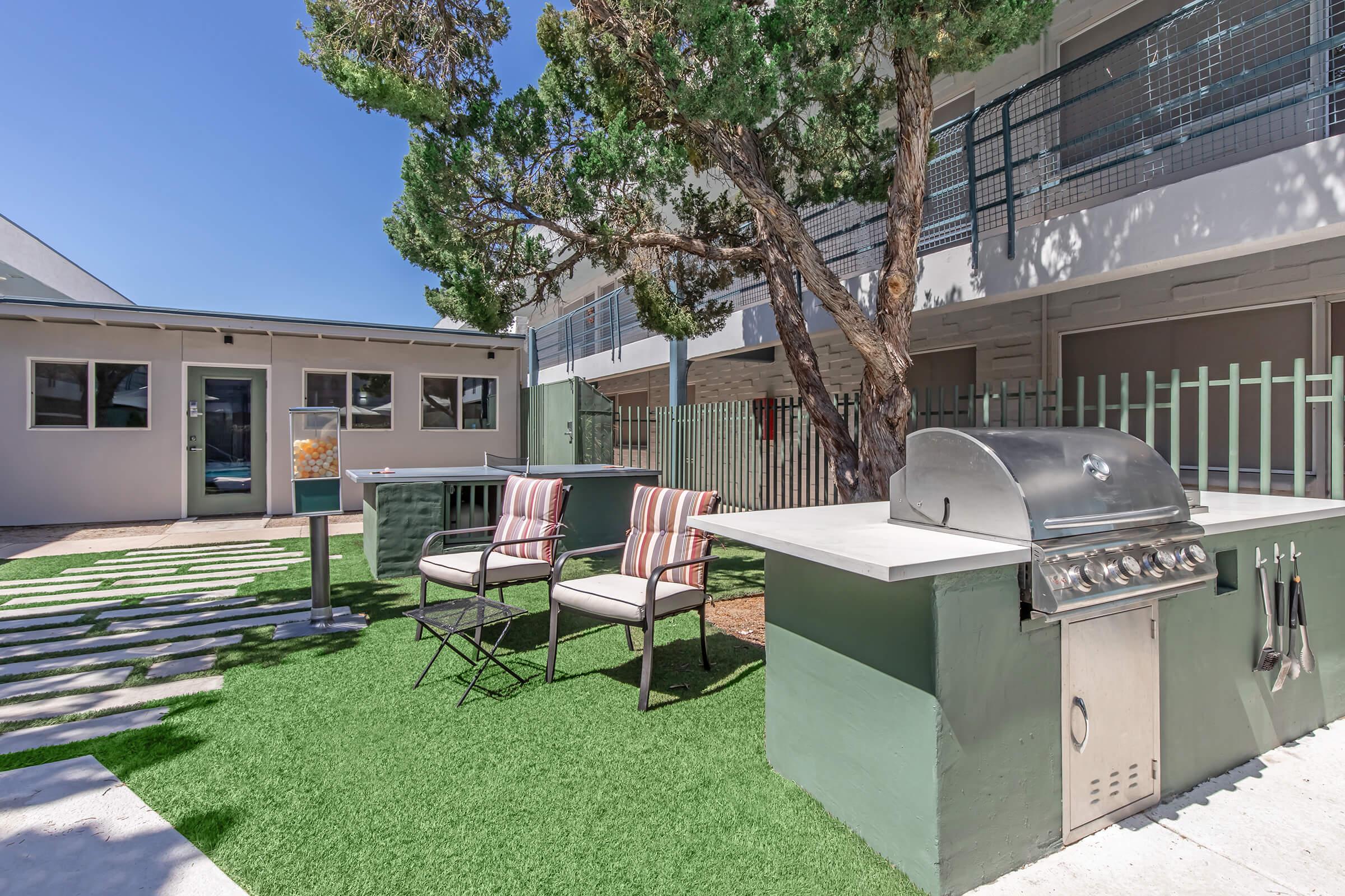 Outdoor space featuring a barbecue grill next to chairs on a lawn, surrounded by greenery. There are two striped chairs and a small table. The backdrop includes a building with several windows and a tree providing shade. Bright blue sky overhead enhances the inviting atmosphere.