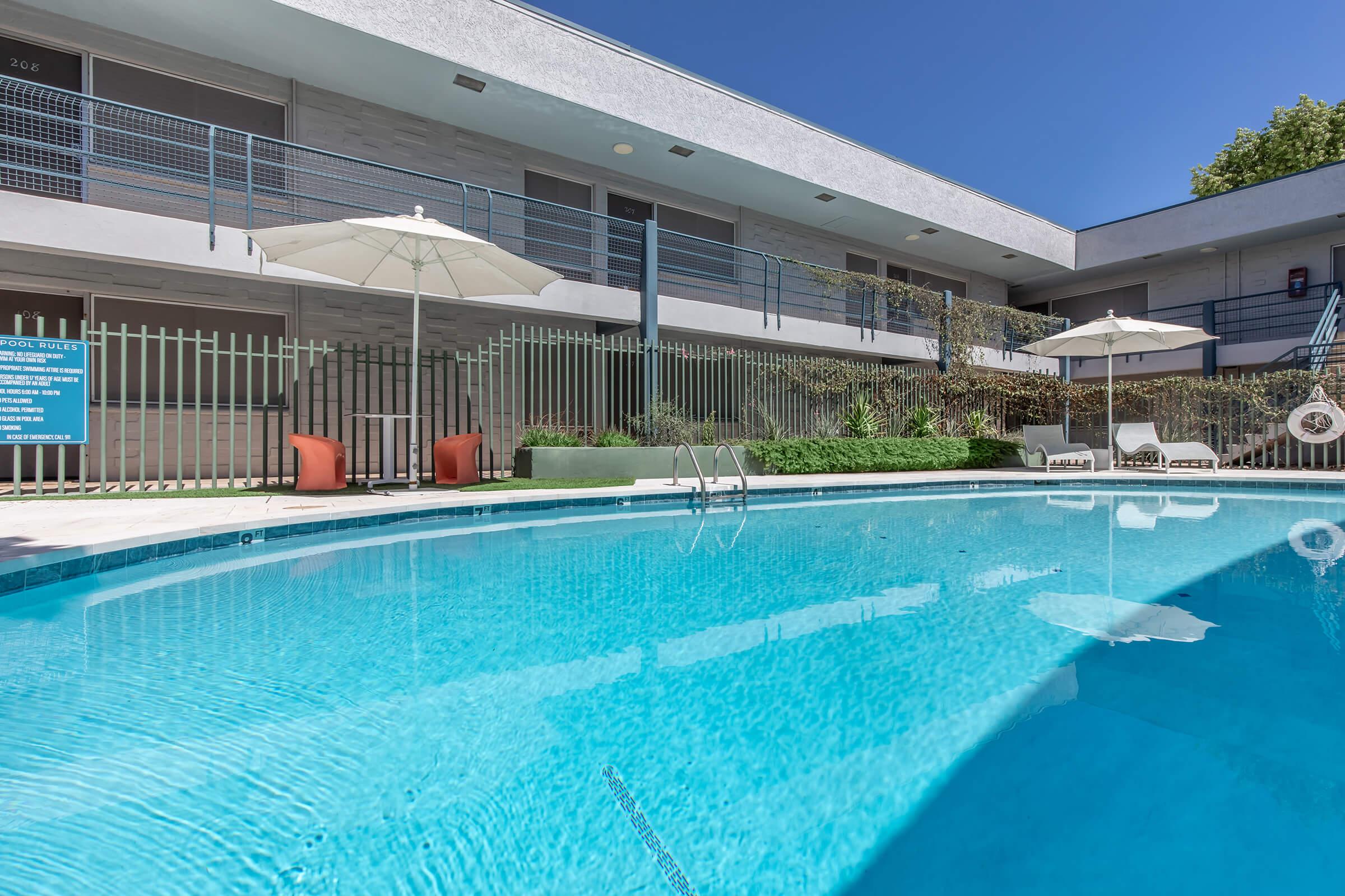 A clear blue swimming pool surrounded by lounge chairs and umbrellas, set against a backdrop of a modern building. The scene is bright and inviting, with greenery along the pool's edge and a bright blue sky overhead.