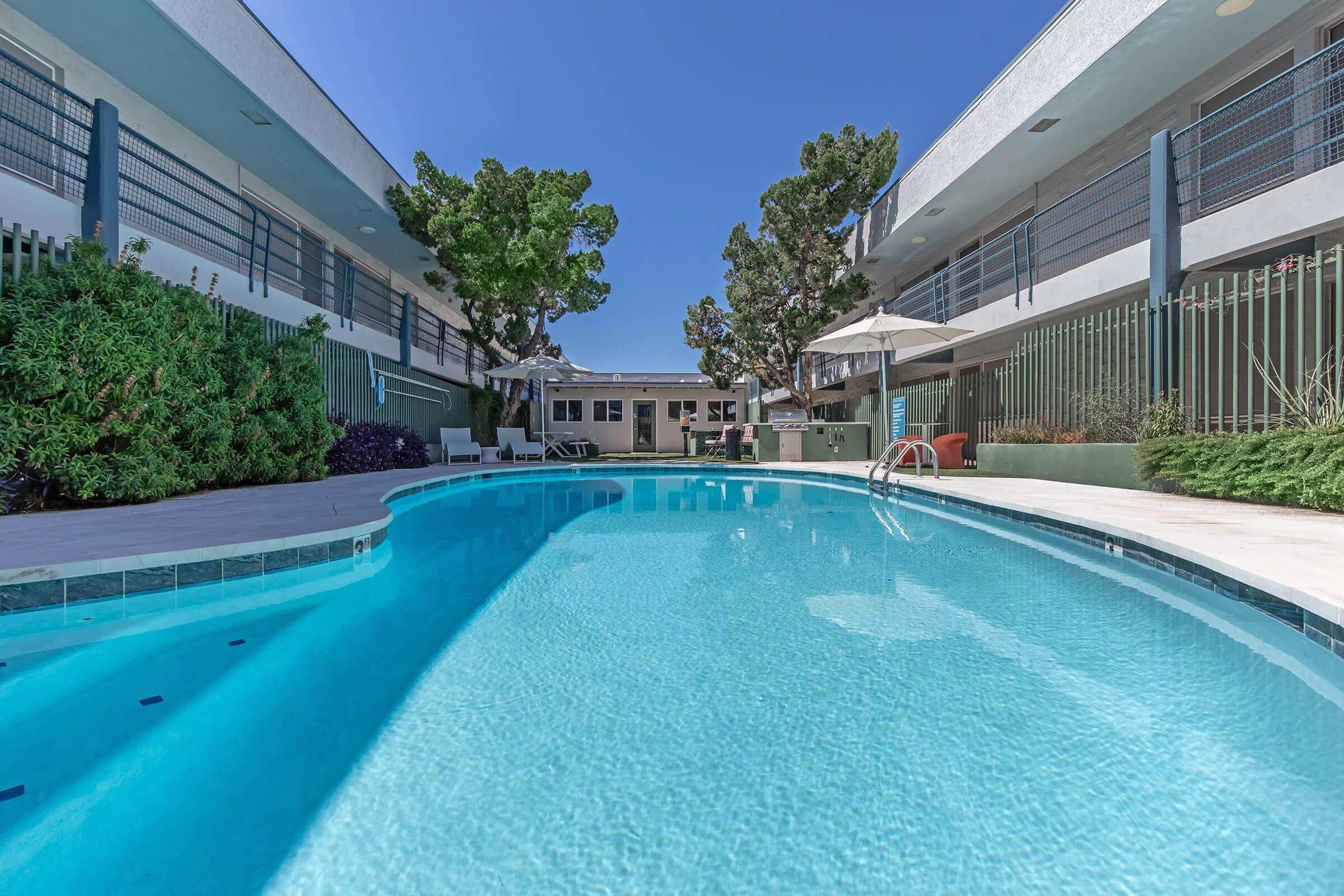 A clear blue swimming pool surrounded by landscaped greenery and modern buildings. Lounge chairs and umbrellas are set up around the pool area, offering a relaxing atmosphere in a sunny environment. The scene conveys a tranquil and inviting setting for leisure activities.