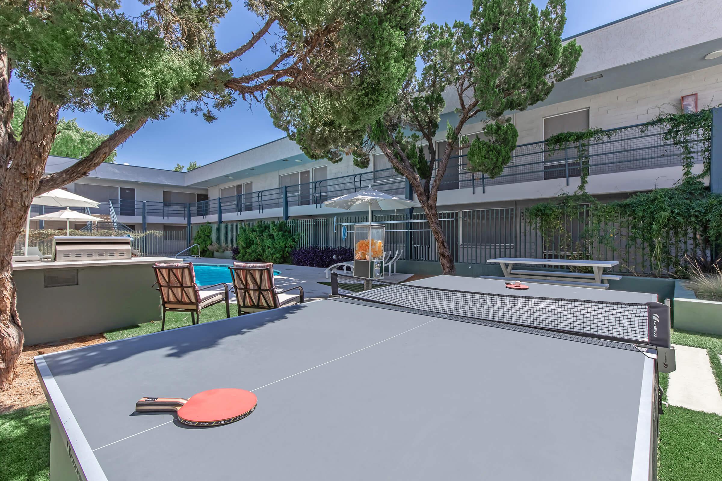 A ping pong table in a sunny courtyard with green trees. In the background, there is a swimming pool, lounge chairs, and patio umbrellas, alongside a modern building. The setting is vibrant and inviting, perfect for outdoor recreation and relaxation.