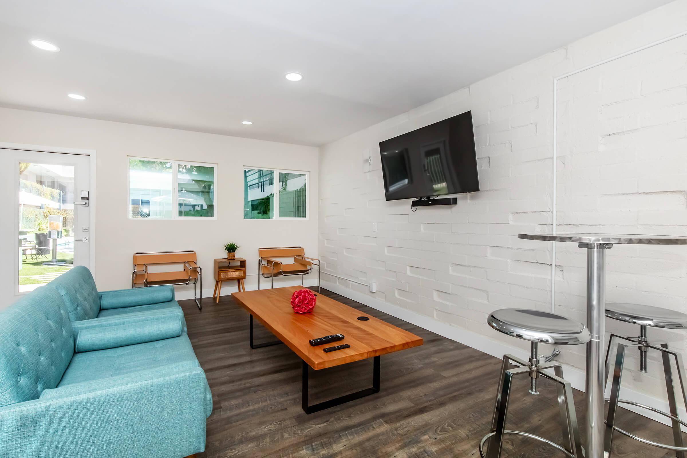 A modern lounge area featuring a light blue couch and two wooden chairs. In the center, there's a wooden coffee table with a decorative red item on it. A flat-screen TV is mounted on the wall, and large windows allow natural light to fill the space. The flooring is a wood-like laminate, creating a warm atmosphere.