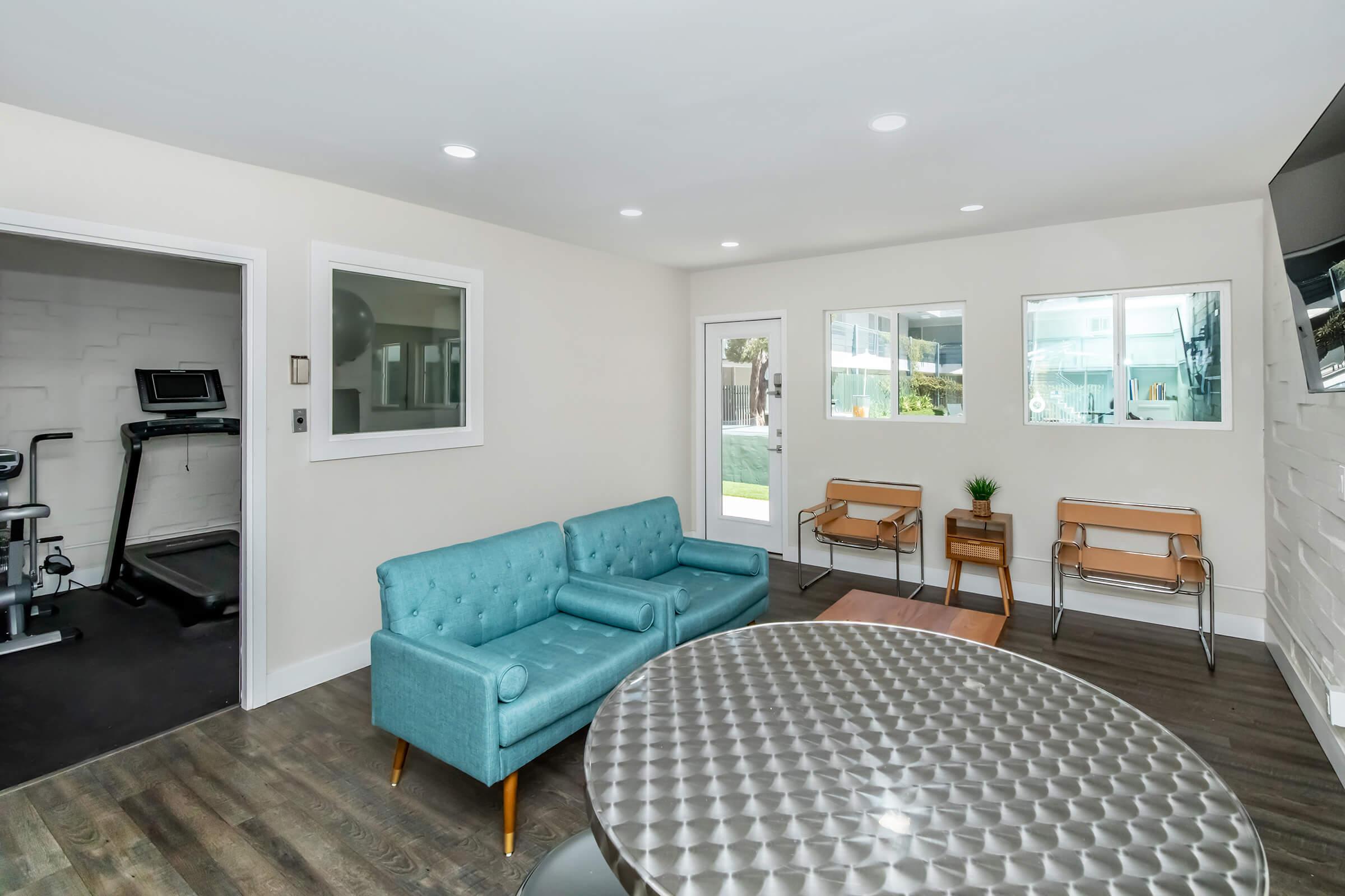 A modern lounge area featuring a light-colored wall, a blue tufted sofa, and a round metallic table. There are two chairs, large windows letting in natural light, and an open doorway leading to an outdoor space. A fitness area is partially visible in the background.