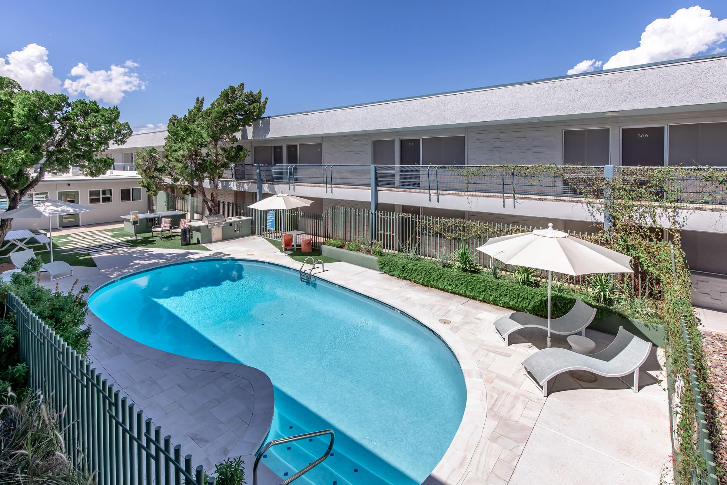 A view of a modern outdoor swimming pool surrounded by lounge chairs and umbrellas, set in a landscaped area. Nearby, a two-story building with balconies overlooks the pool area under a clear blue sky with a few clouds. The scene conveys a relaxed, resort-like atmosphere.
