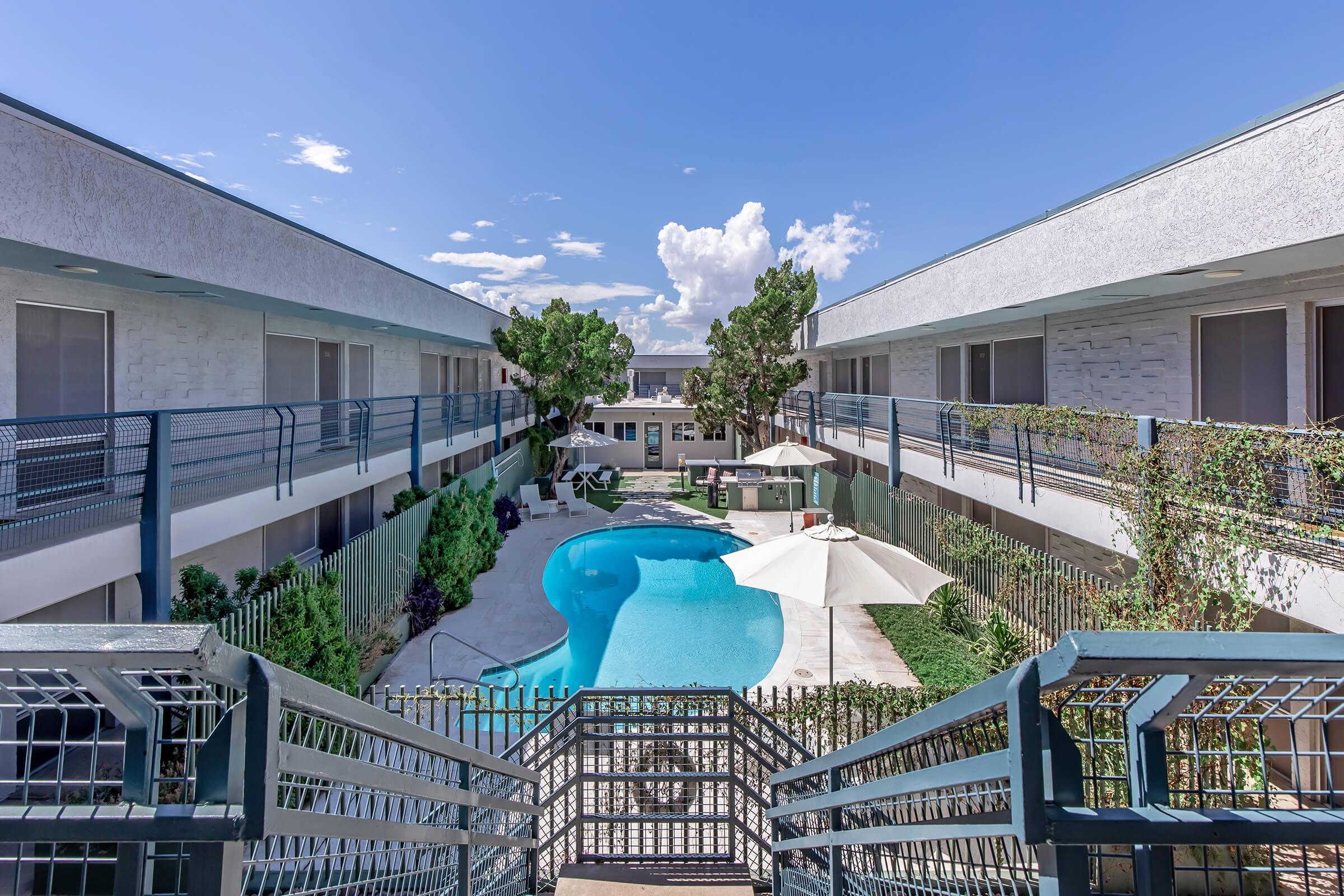 A scenic view of an apartment complex featuring a swimming pool surrounded by well-maintained landscaping. Two buildings flank the pool area, and there are lounge chairs and umbrellas for relaxation. Bright blue sky with a few clouds enhances the inviting atmosphere of the space.