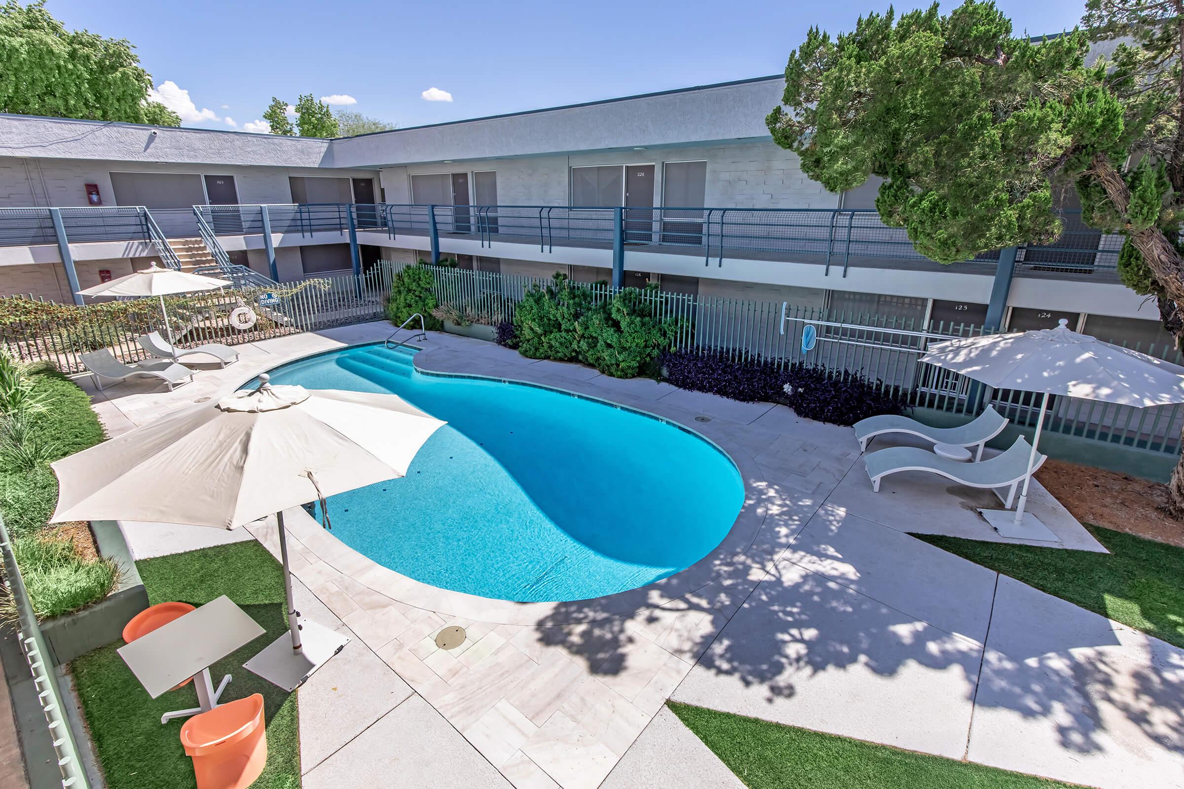 A bright, tranquil pool area surrounded by lounge chairs and umbrellas, located near a multi-story building. Lush greenery and a clear blue sky enhance the inviting atmosphere. The pool's curved shape adds a modern touch to the outdoor space, perfect for relaxation and leisure.