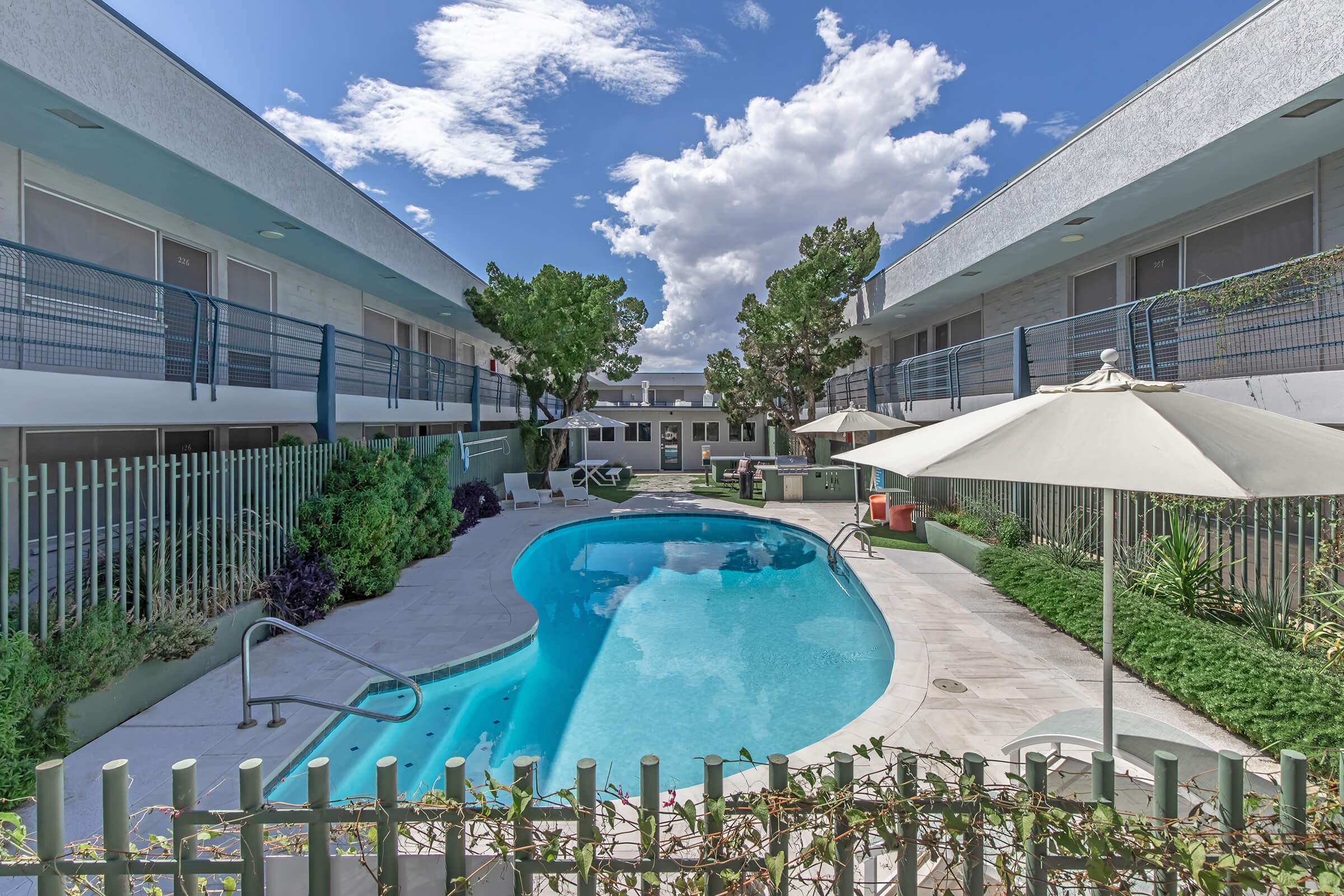 A bright and inviting swimming pool surrounded by greenery, with patio chairs and umbrellas. The pool is located in a courtyard between two buildings. The sky is partly cloudy, adding a serene ambiance to the scene.