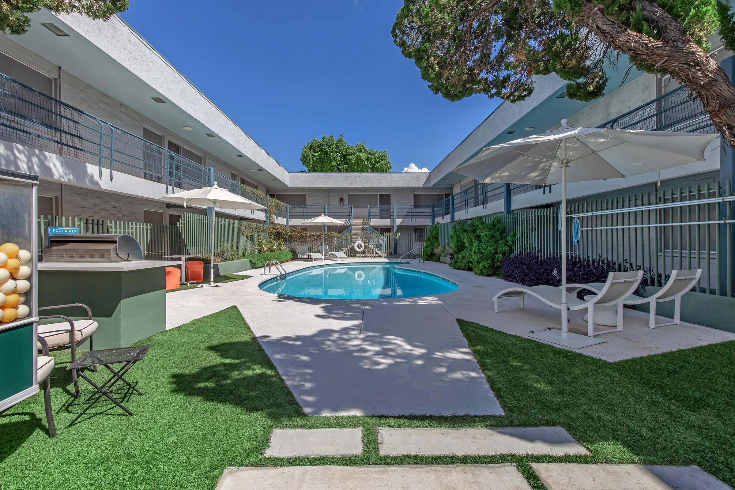 A sunny courtyard featuring a swimming pool surrounded by lounge chairs, umbrellas, and lush greenery. The area includes an outdoor seating space and a storage unit with a display of colorful items, all set against a clear blue sky.