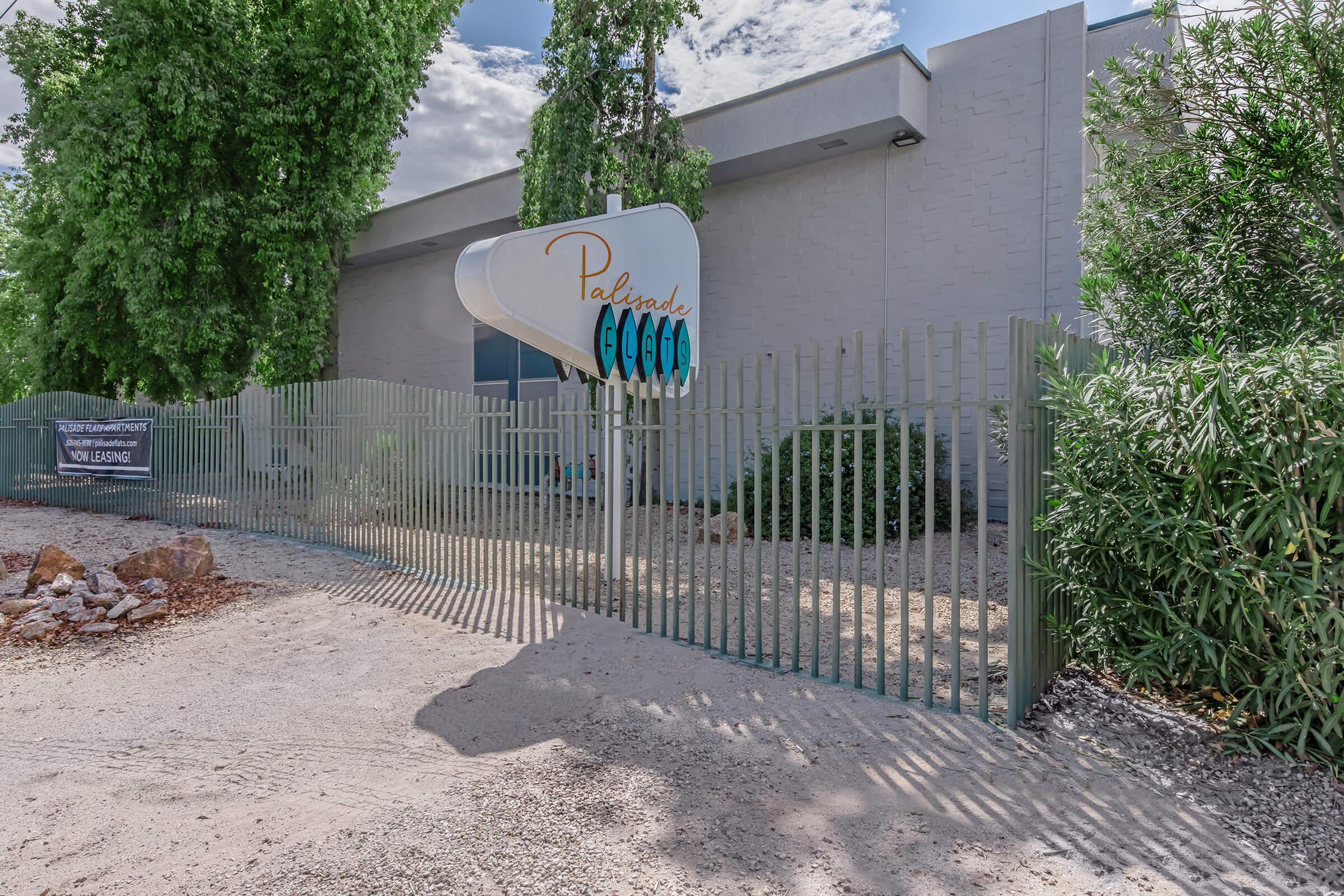Exterior view of a building with a prominent sign that reads "Palms." The entrance is enclosed by a green fence, and there are surrounding trees and shrubs. A banner displaying "Now Leasing" is visible on the fence, set against a clear blue sky with some clouds. The ground is a mix of gravel and dirt.