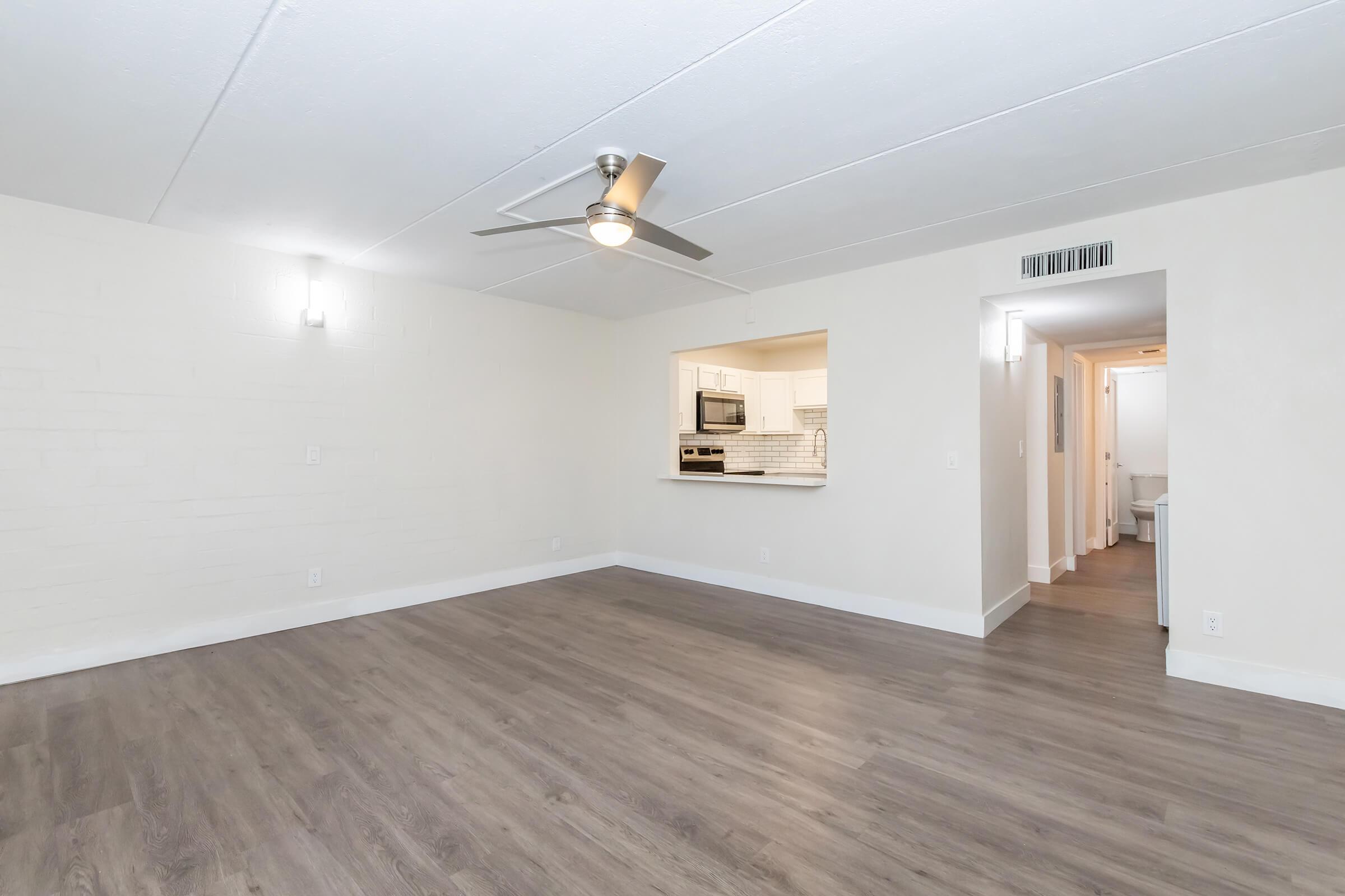 Spacious and bright living area with light-colored walls and modern flooring. Features a ceiling fan and an open layout connecting to a kitchen area. Doorways lead to other rooms, creating an inviting atmosphere in a contemporary indoor space.