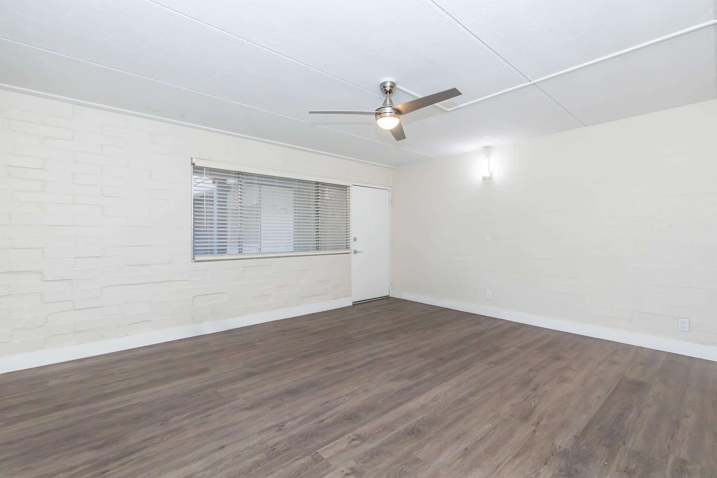 Empty room with light-colored walls and a ceiling fan. It features a window with blinds and a door leading outside. The flooring is a warm wooden laminate. The space is well-lit, creating an open and airy atmosphere.