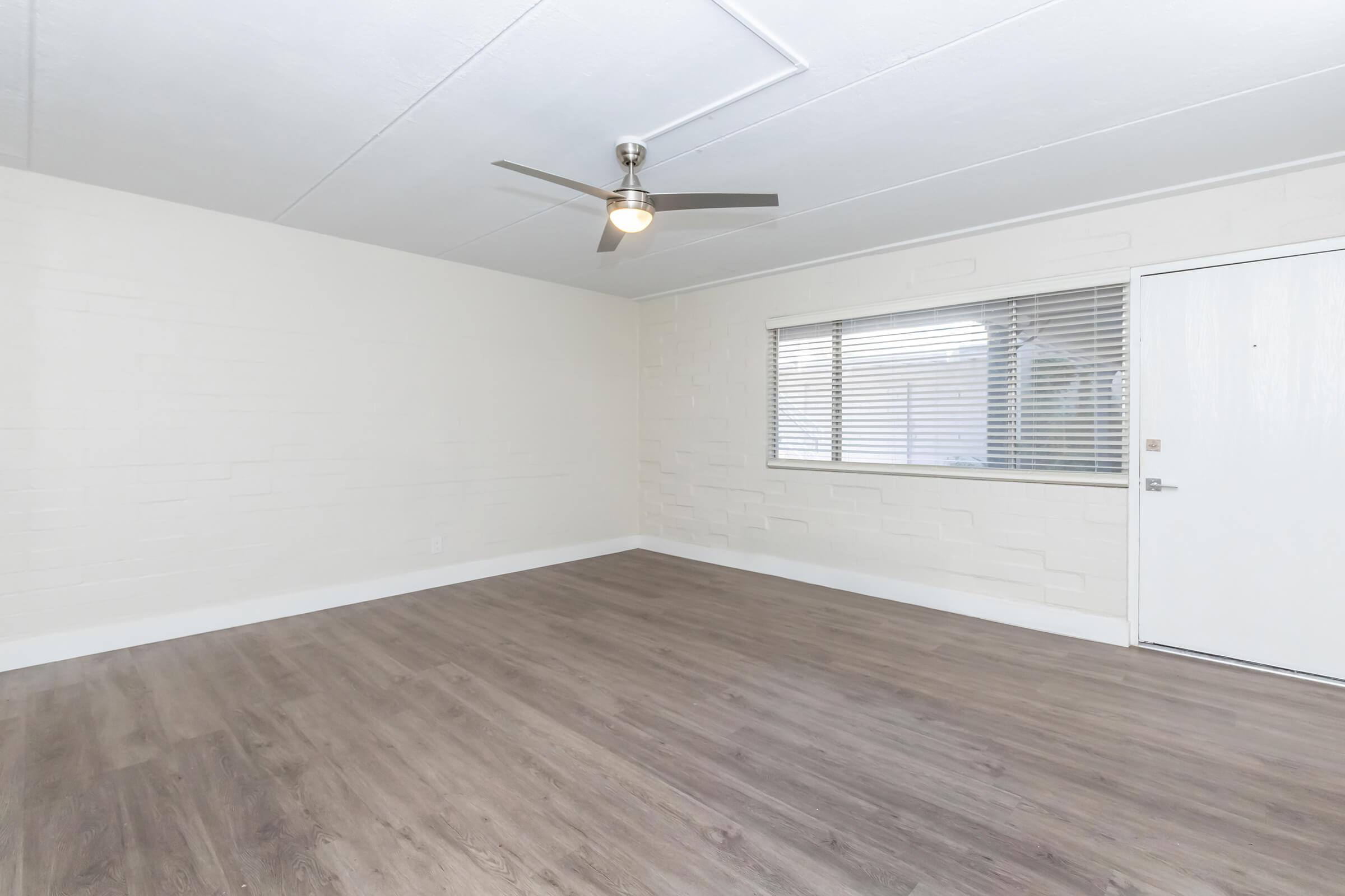 Interior view of a vacant room with light-colored walls and a ceiling fan. The flooring is a light wood laminate. A window allows natural light into the space, and there is a white door visible on the right side. The room is empty and appears freshly painted, suitable for various uses or furnishing.
