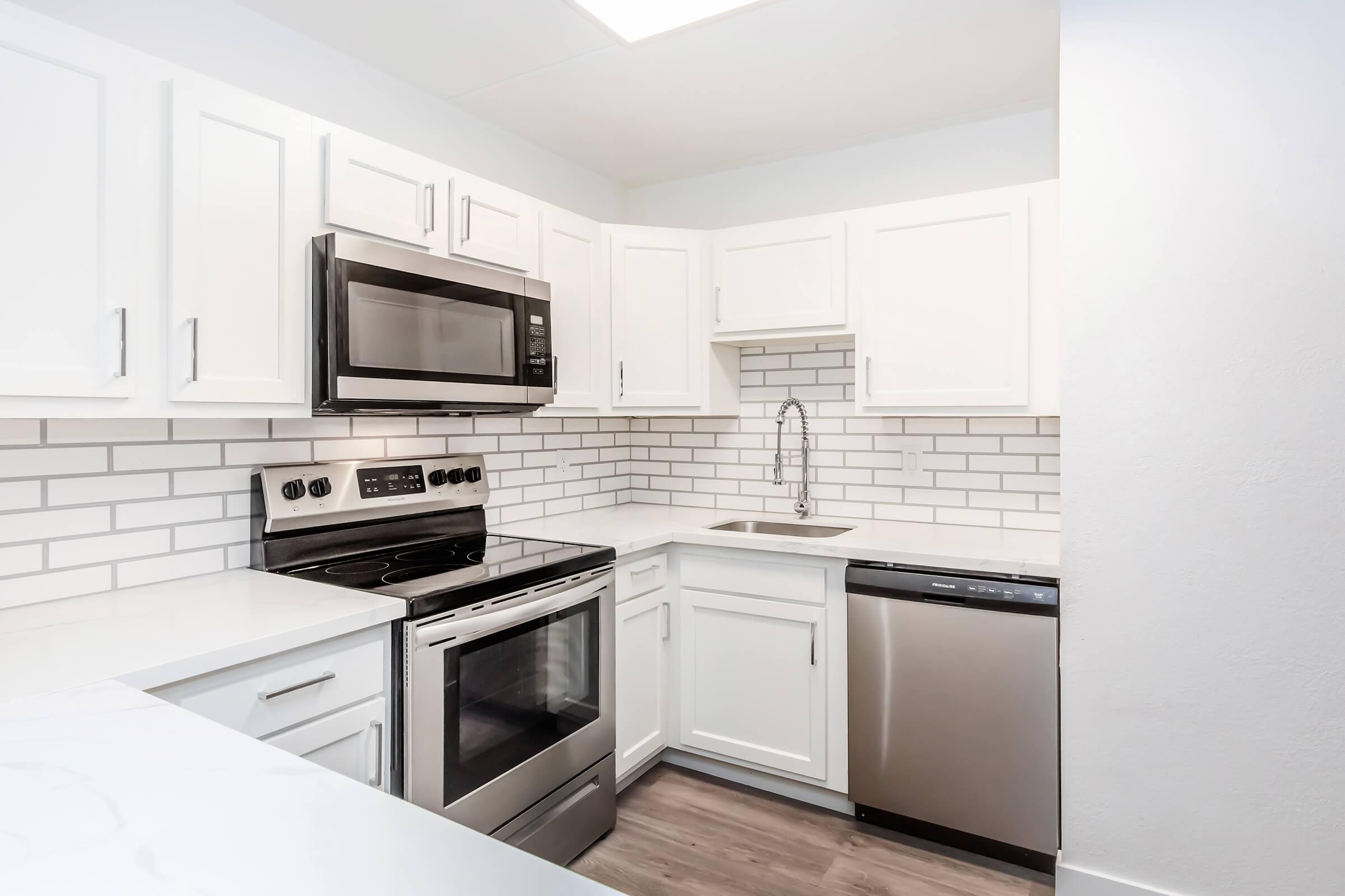 Modern kitchen featuring white cabinets, stainless steel appliances including a microwave, oven, and dishwasher, a sleek countertop, and a tiled backsplash. Light, neutral colors create a bright and clean aesthetic with ample workspace.