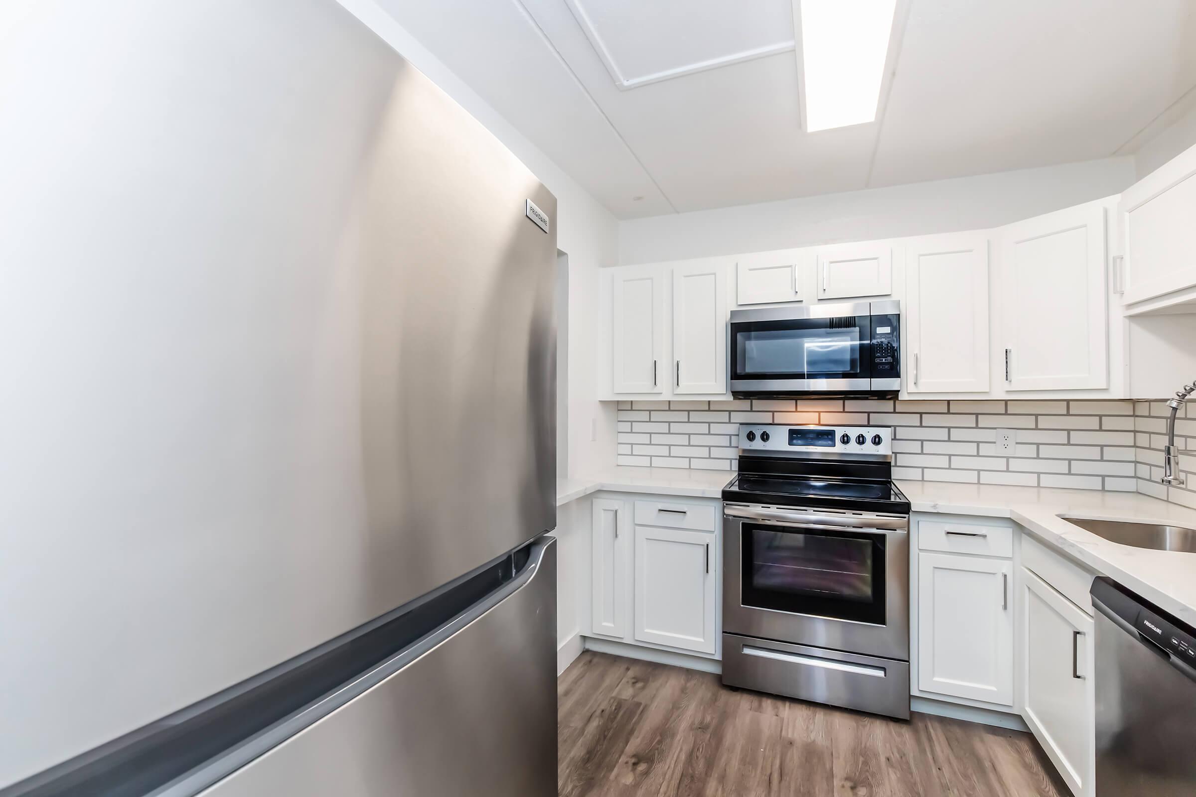 Modern kitchen featuring stainless steel appliances, including a refrigerator and an oven with a microwave above. The cabinets are white, complemented by a light-colored tiled backsplash. The flooring is a wood-like design, and there is ample natural light from a ceiling fixture.